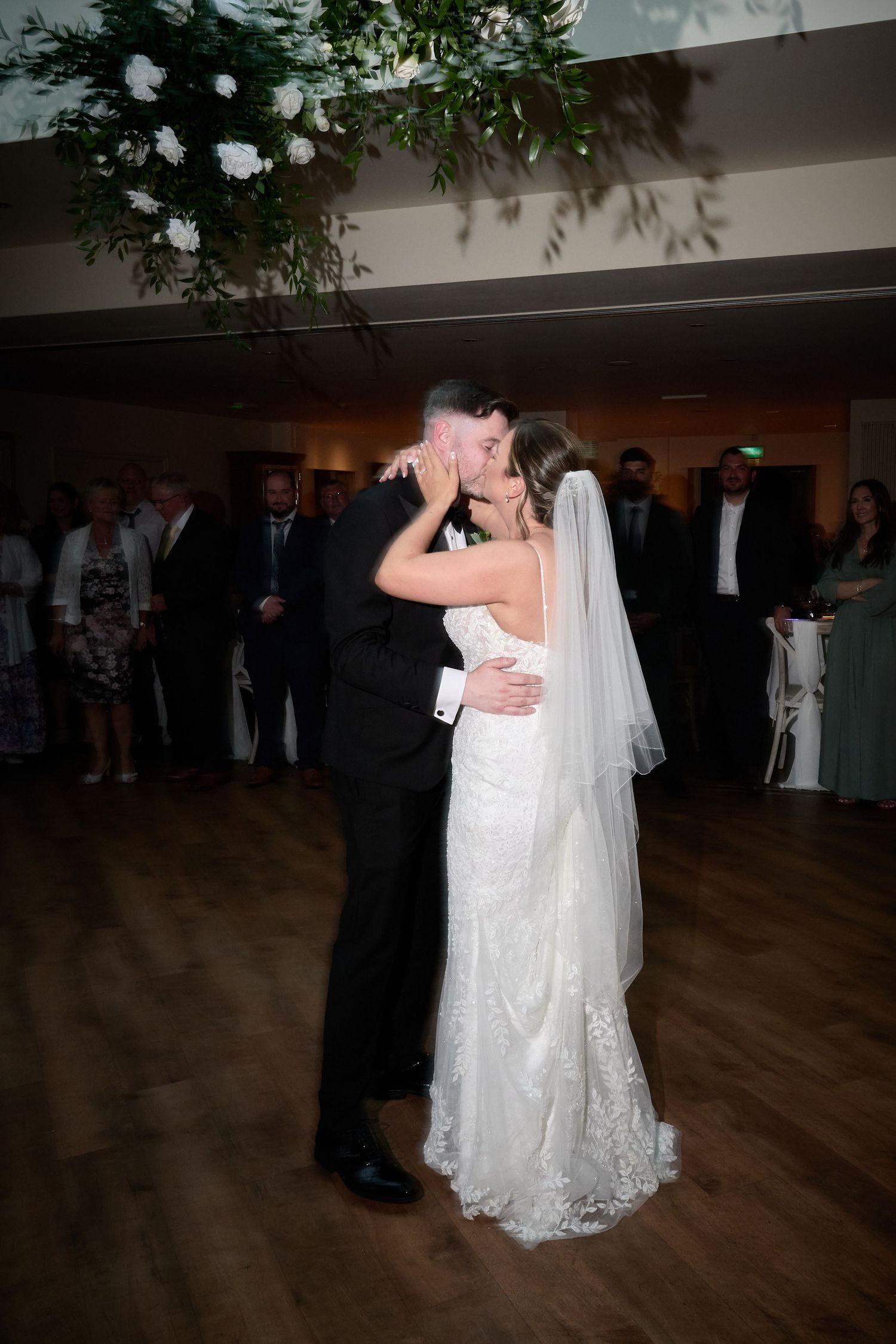 Bride and groom share first dance kiss in elegant wedding reception venue with floral ceiling installation.