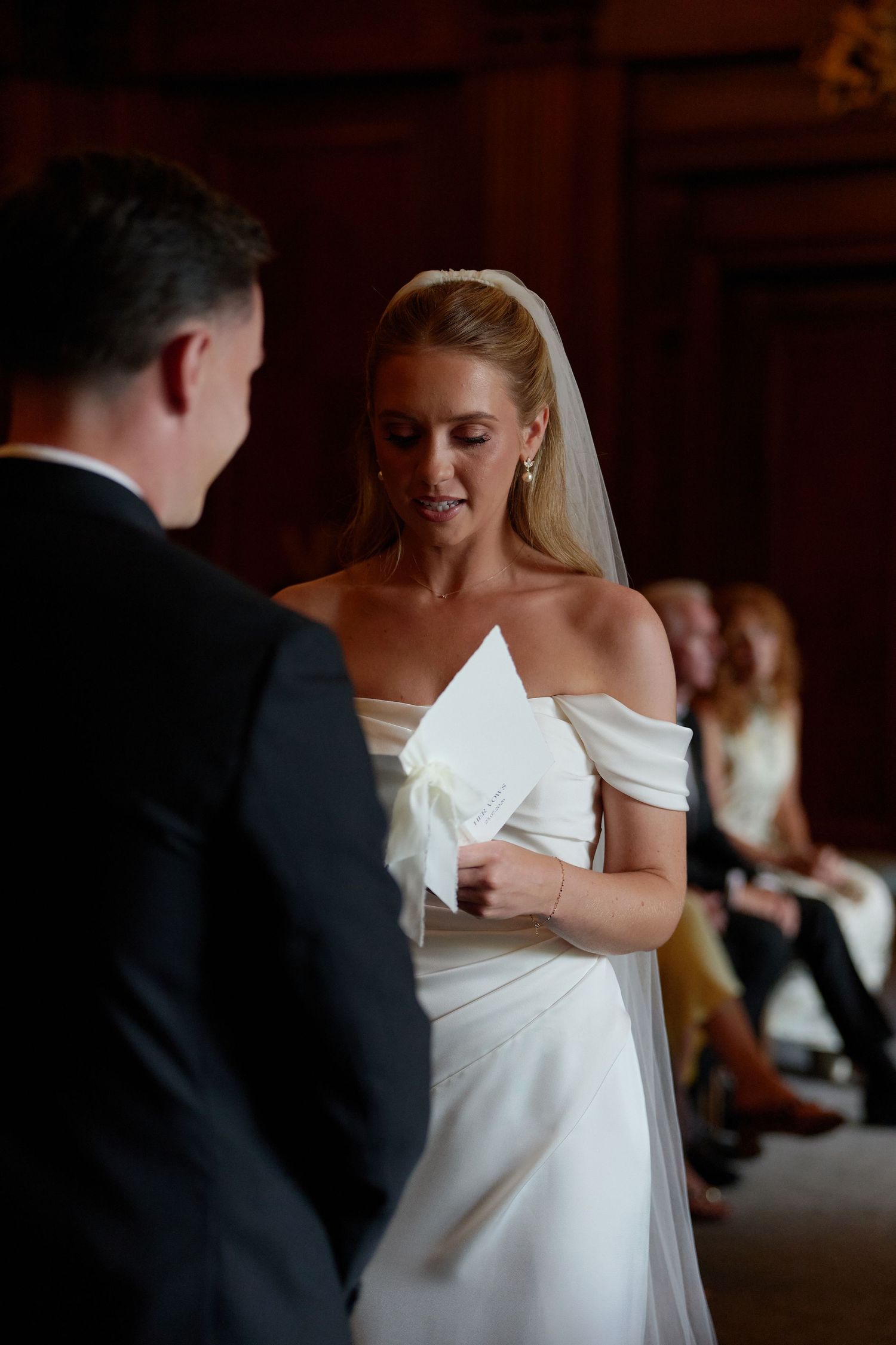 Bride in off-shoulder white gown reads vows during wedding ceremony, wearing veil and pearl earrings.