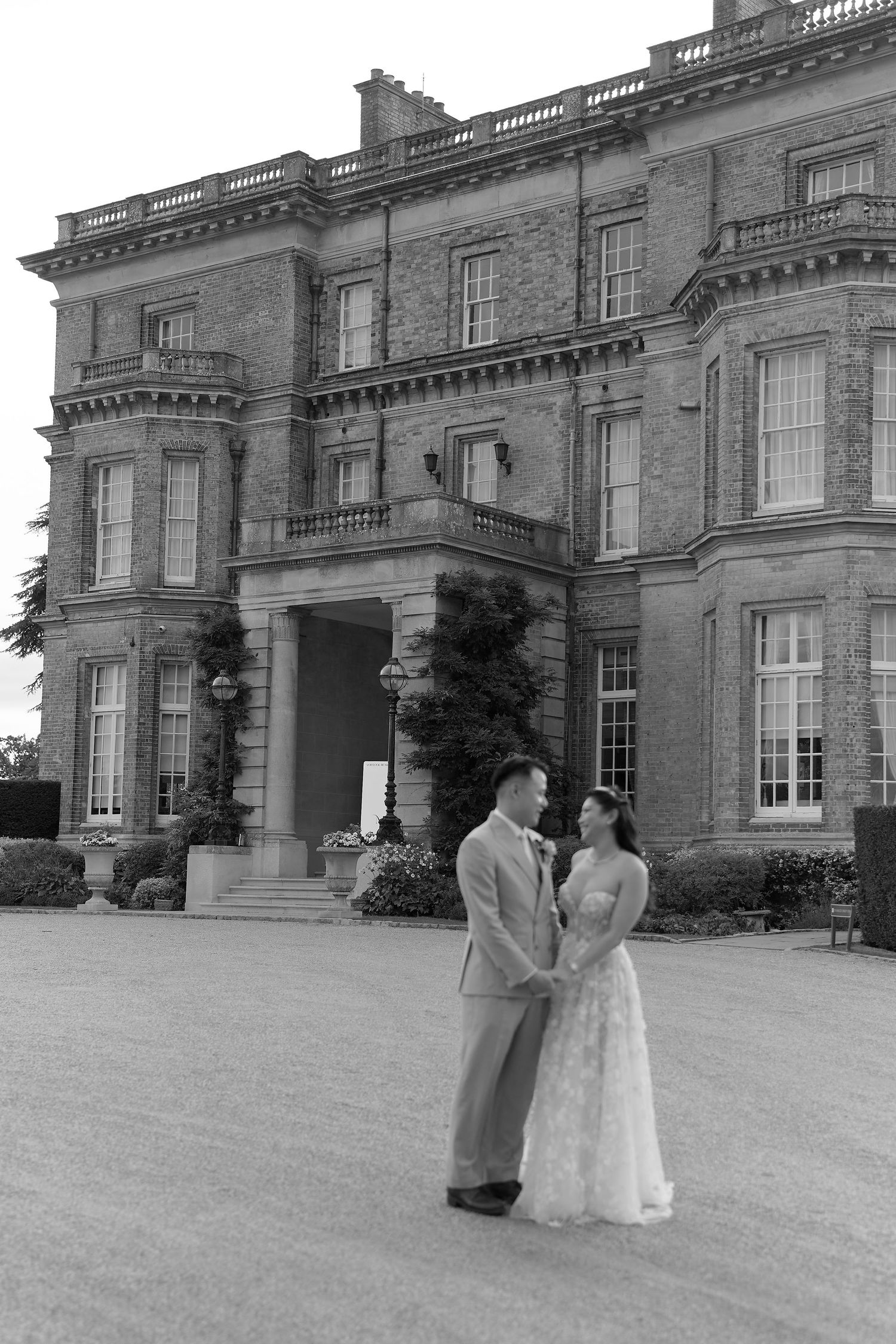 Black and white photo of a couple in wedding attire holding hands before a grand historic brick manor house.