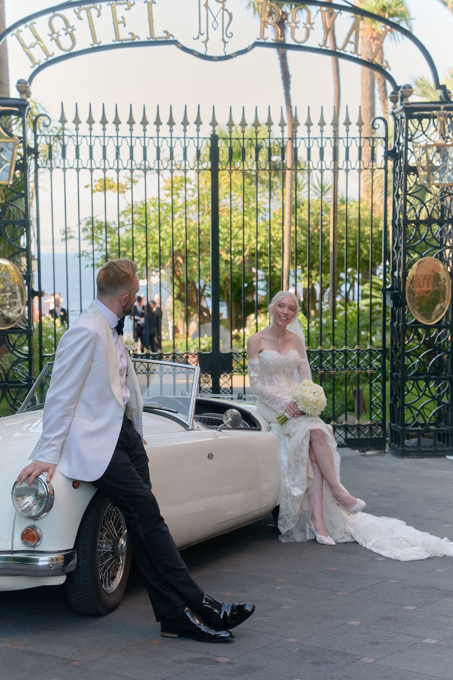 Bride and groom pose by a vintage white convertible at Hotel Royal gates, bride holding white bouquet.
