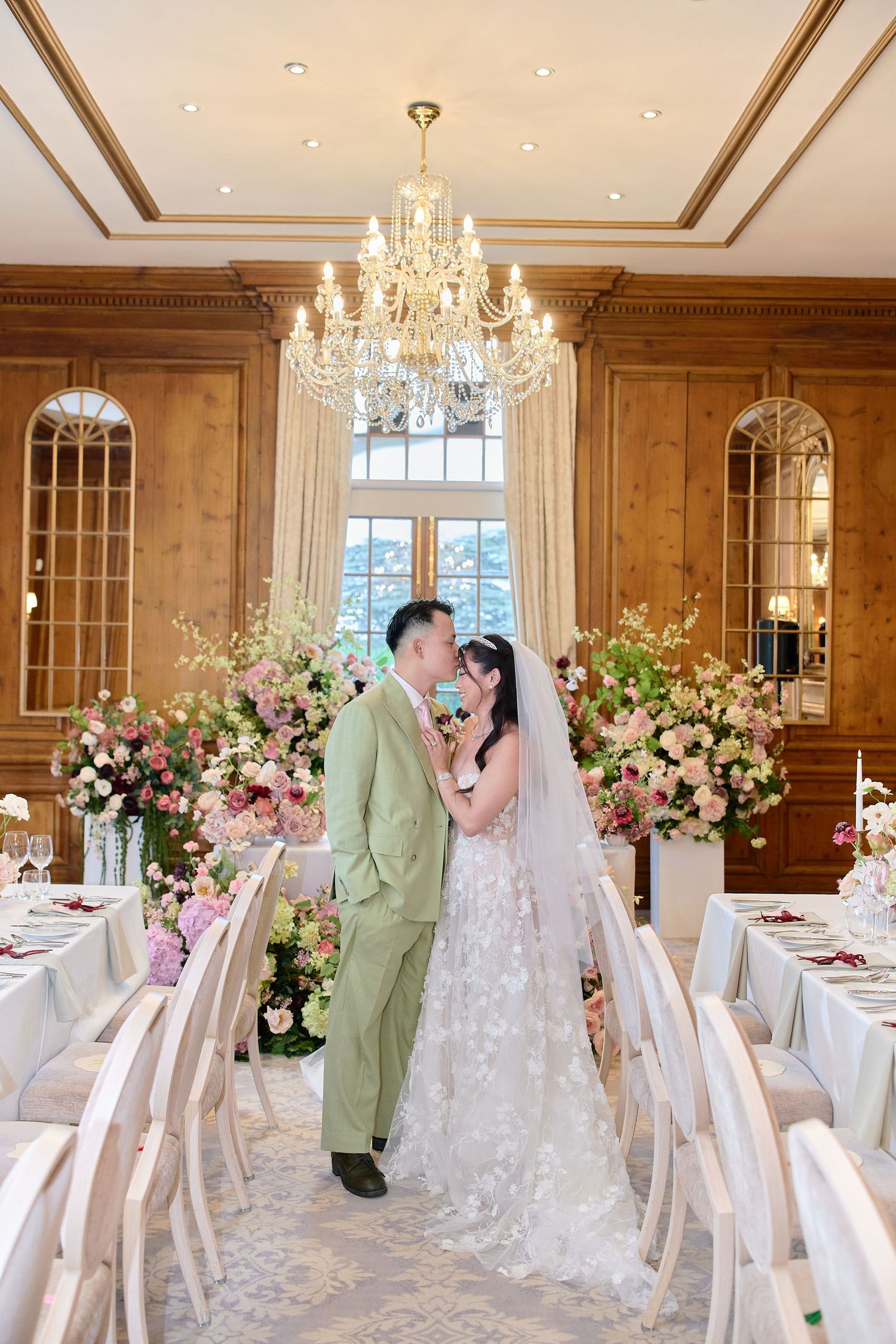 Bride and groom share a kiss in an elegant wedding reception room with pink floral arrangements and a crystal chandelier.