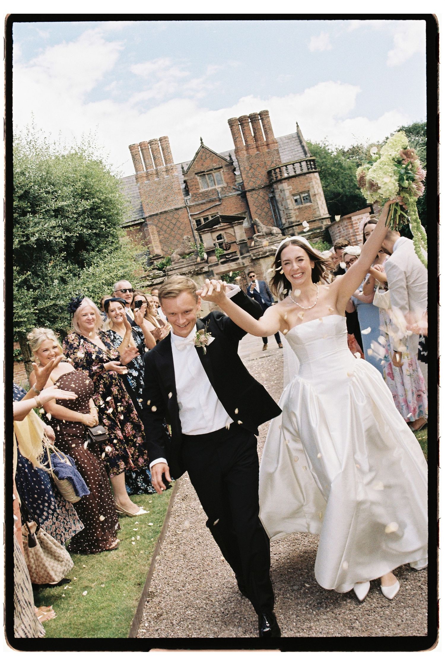 Joyful newlyweds walk through confetti shower outside historic English manor surrounded by cheering wedding guests.