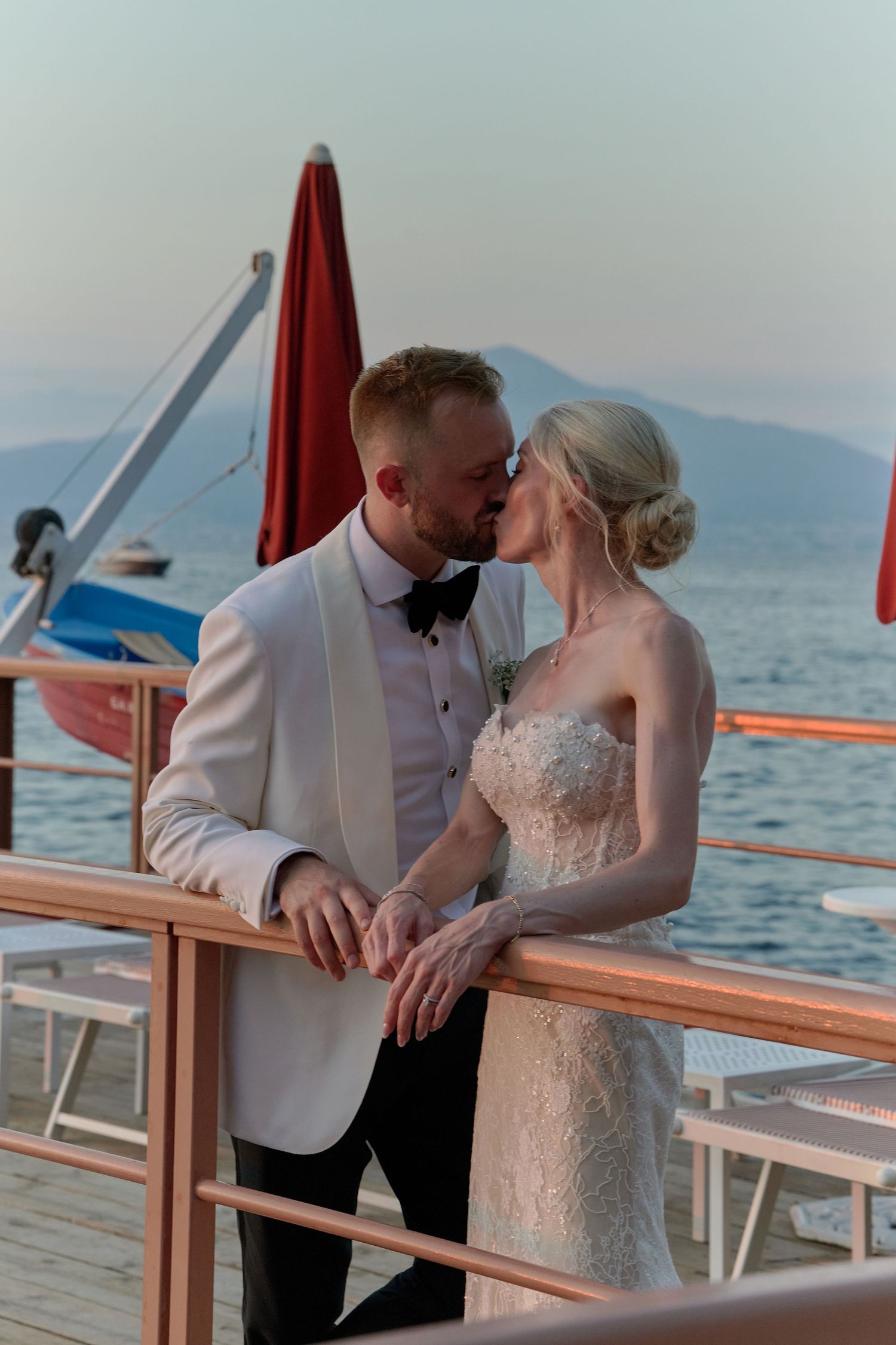 Bride and groom kissing on a boat deck with sea and mountains in background at sunset.