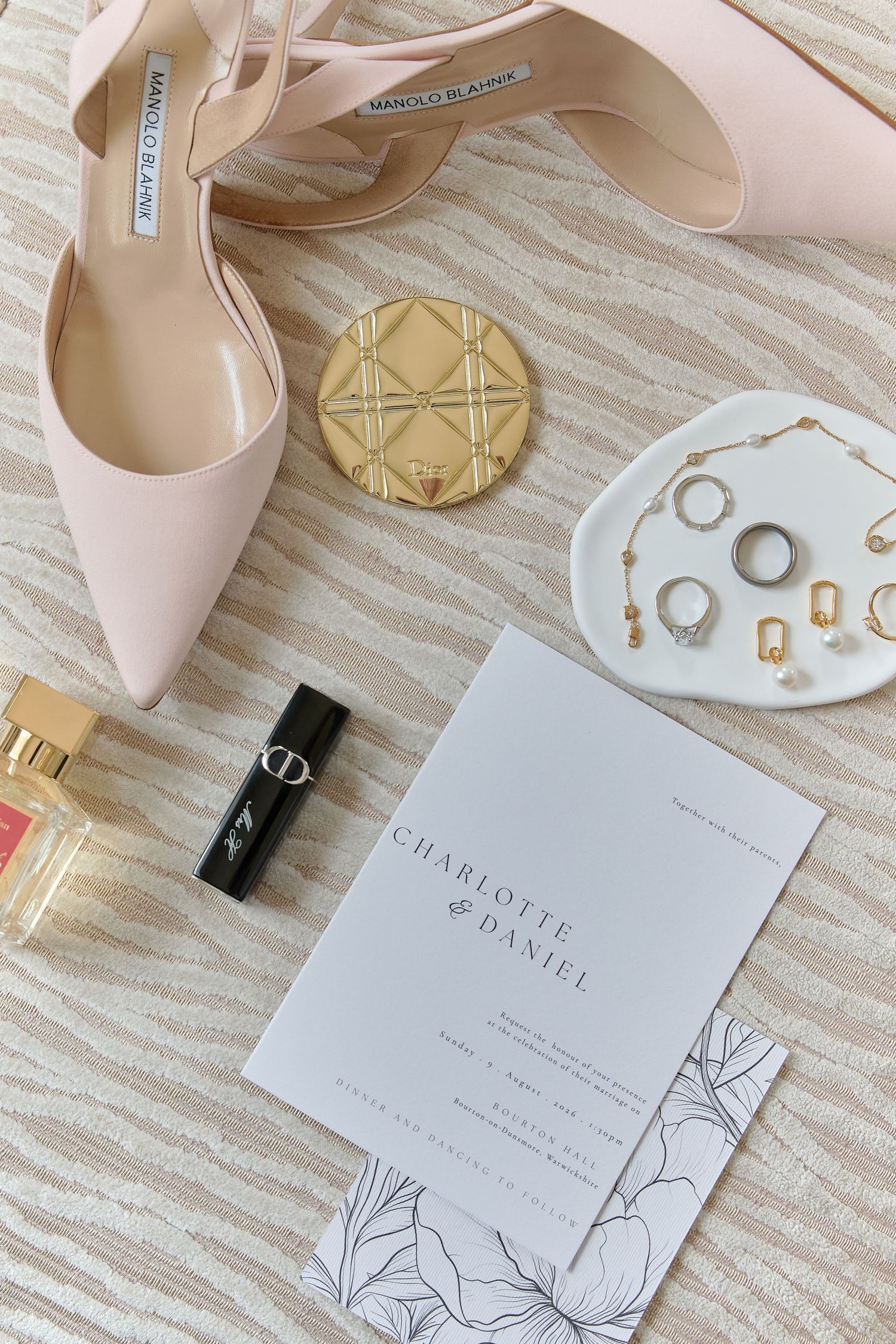 Elegant wedding flat lay with Manolo Blahnik heels, gold compact, jewelry, perfume, and Charlotte & Daniel invitation.