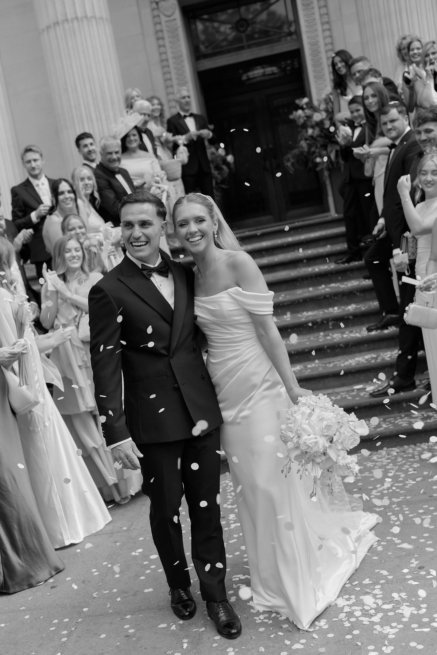 Newlyweds smile as guests throw confetti outside elegant venue steps in black and white photo.