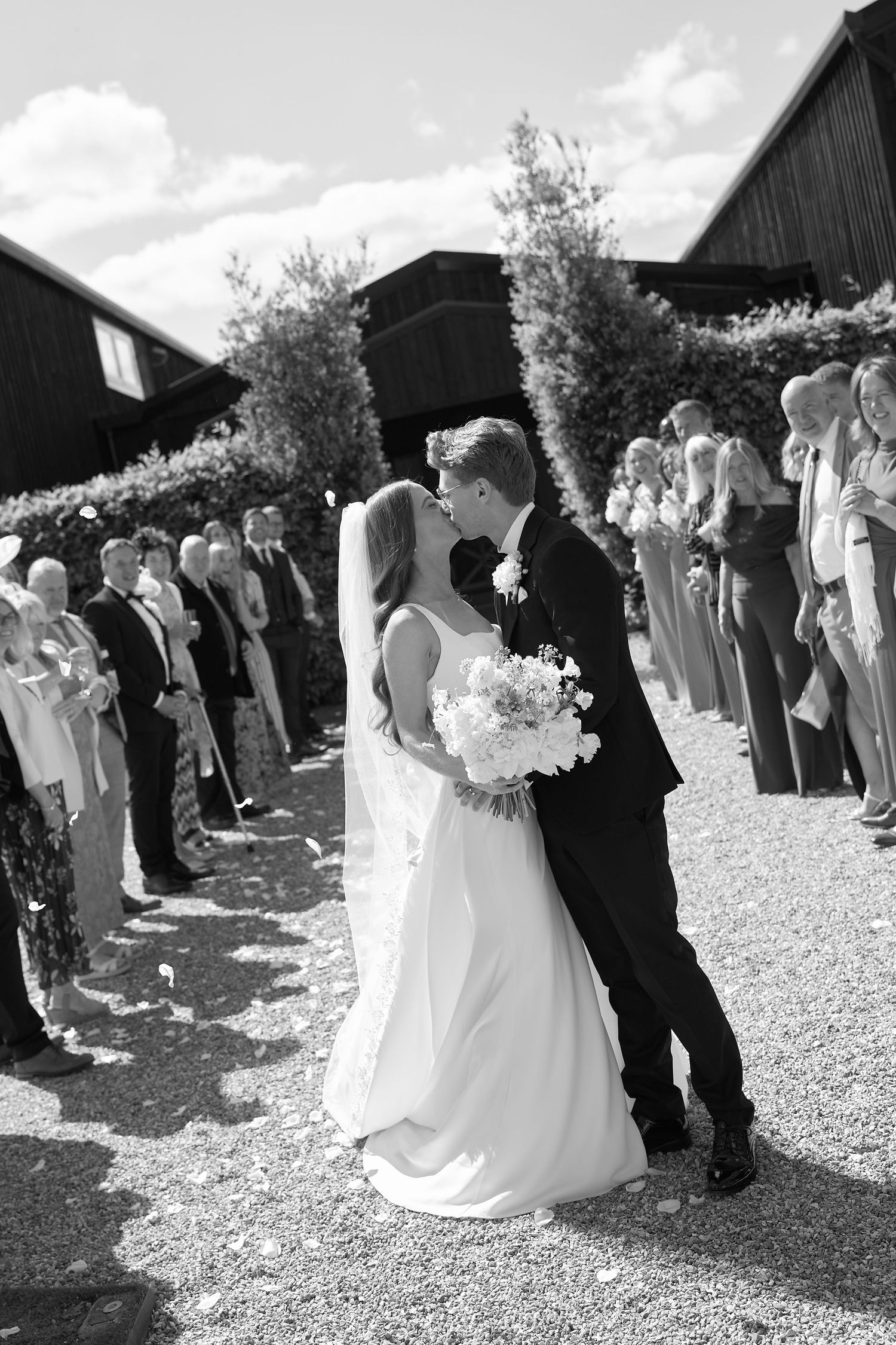 Bride and groom share first kiss as newlyweds while guests line the aisle in black and white.
