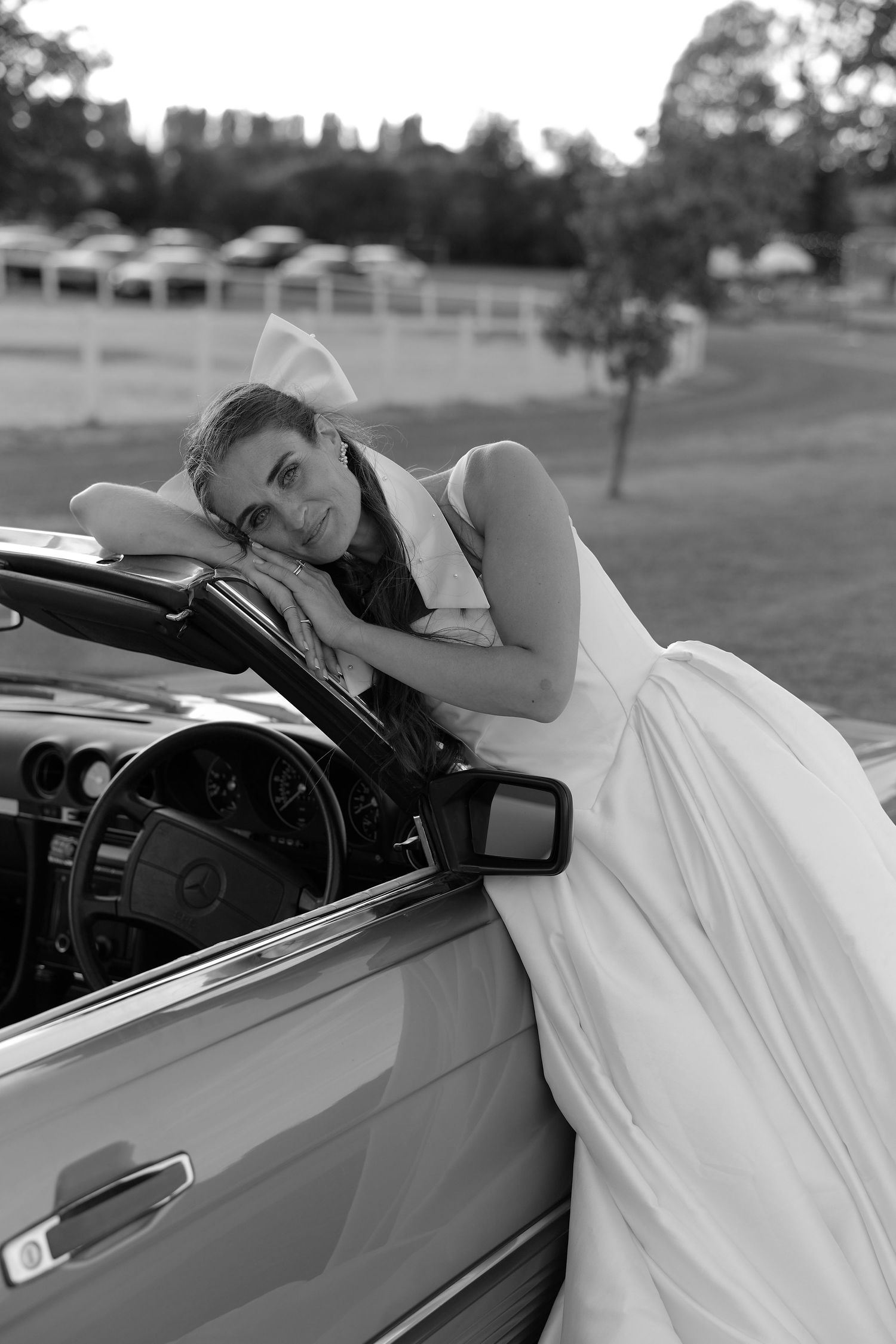Black and white photo of a bride in a white gown leaning on a vintage convertible.