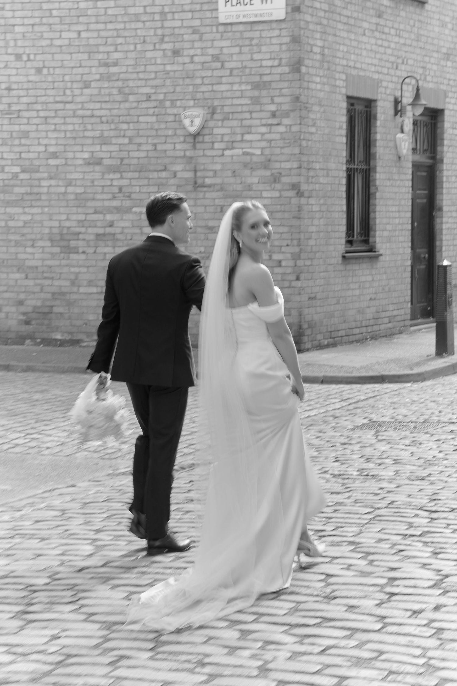 Black and white photo of a smiling bride in white gown and groom in suit walking on cobblestone street.