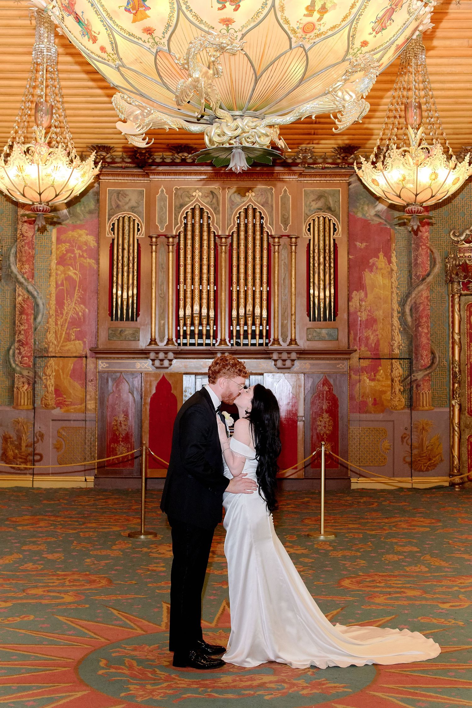 Bride and groom share a romantic kiss in an ornate ballroom with golden chandeliers and decorative pipe organ backdrop.