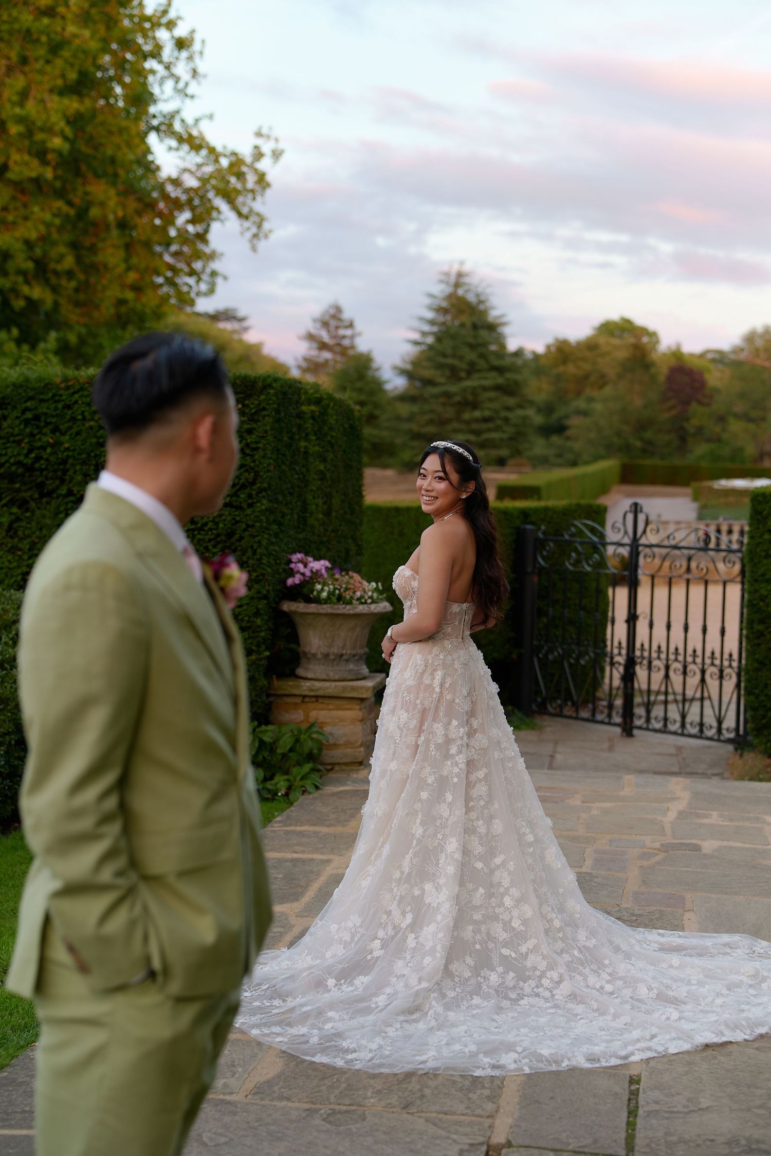 Bride in floral lace gown smiles at groom in sage suit during golden hour garden wedding portrait.
