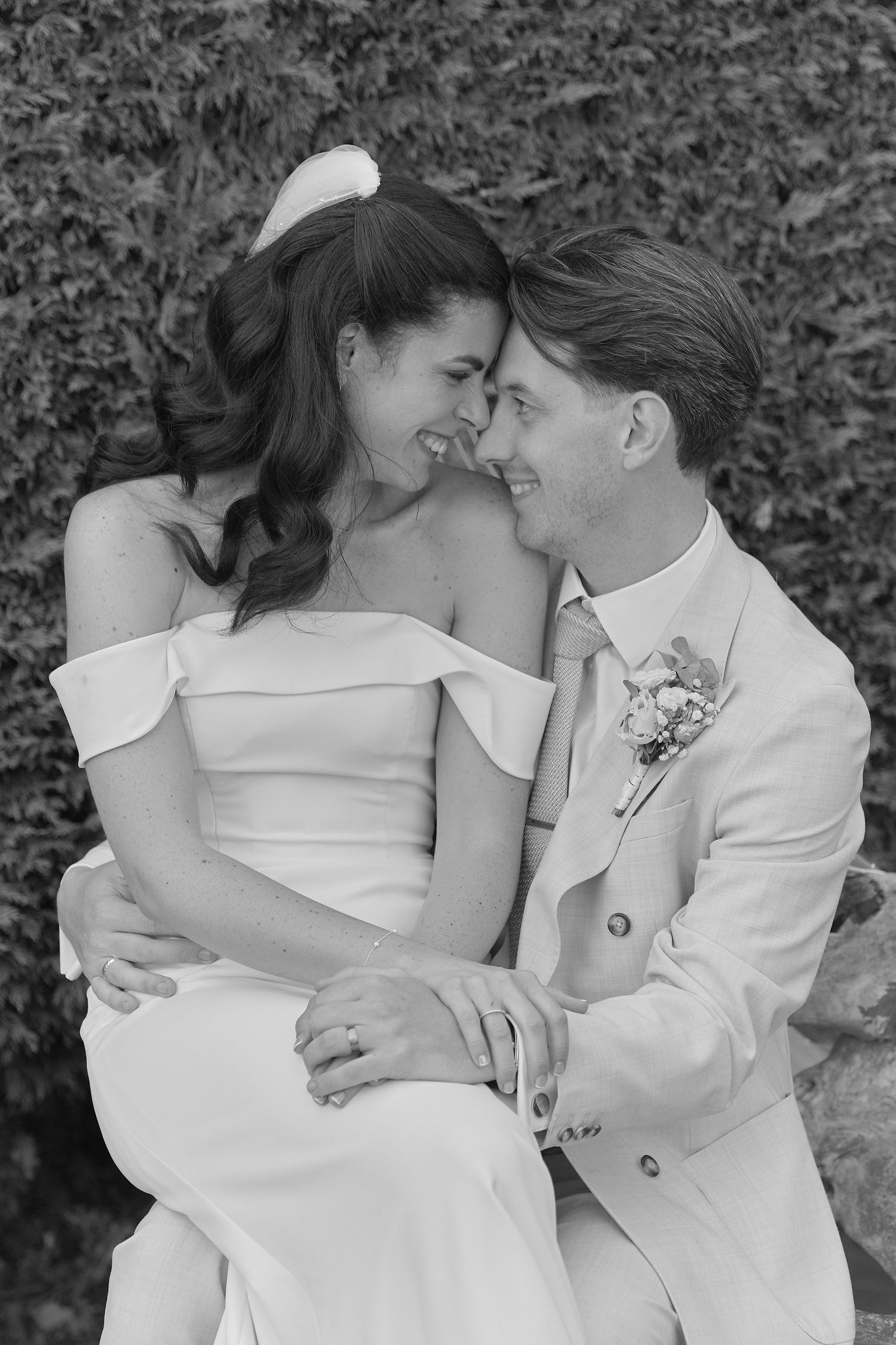 Black and white photo of a smiling bride and groom in wedding attire, sitting closely together and gazing at each other.