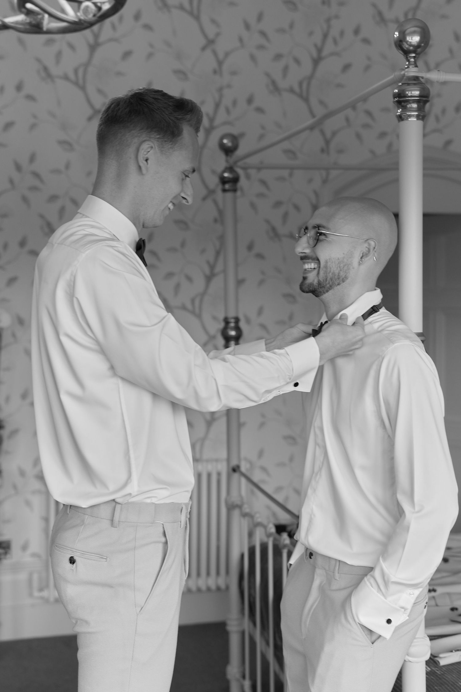 Black and white photo of two grooms smiling and adjusting bow ties while getting ready for their wedding.