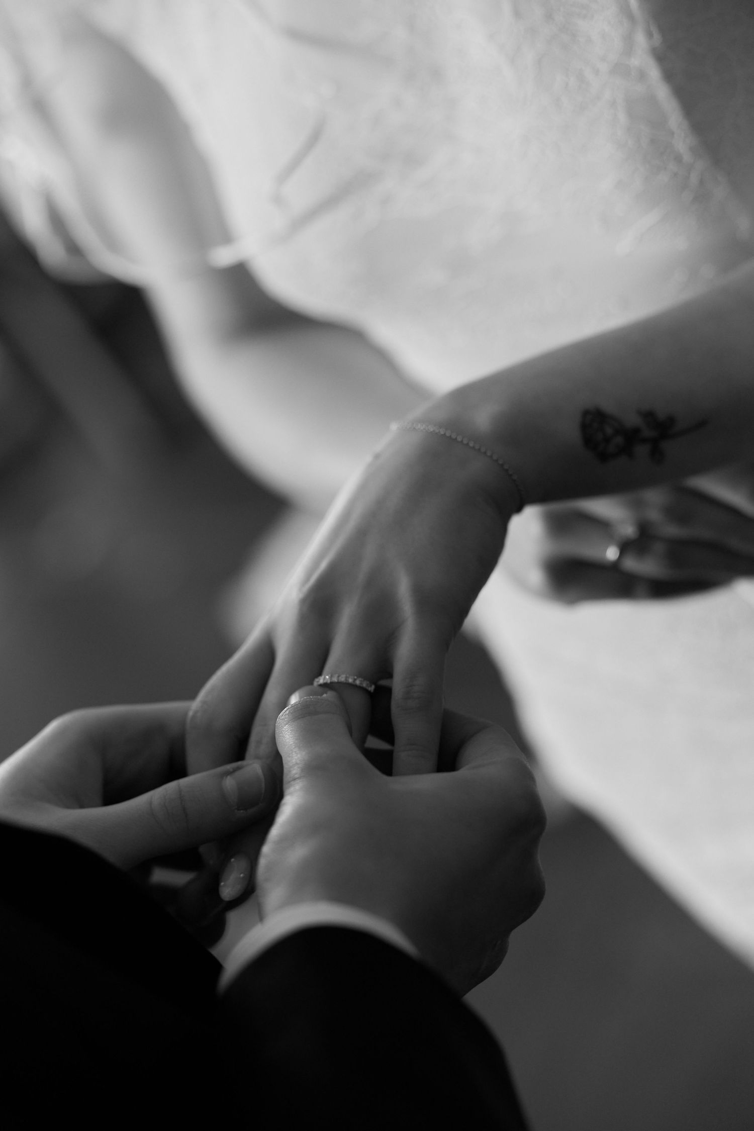 Black and white photo of groom placing wedding ring on bride's finger, with small butterfly tattoo visible on her wrist.