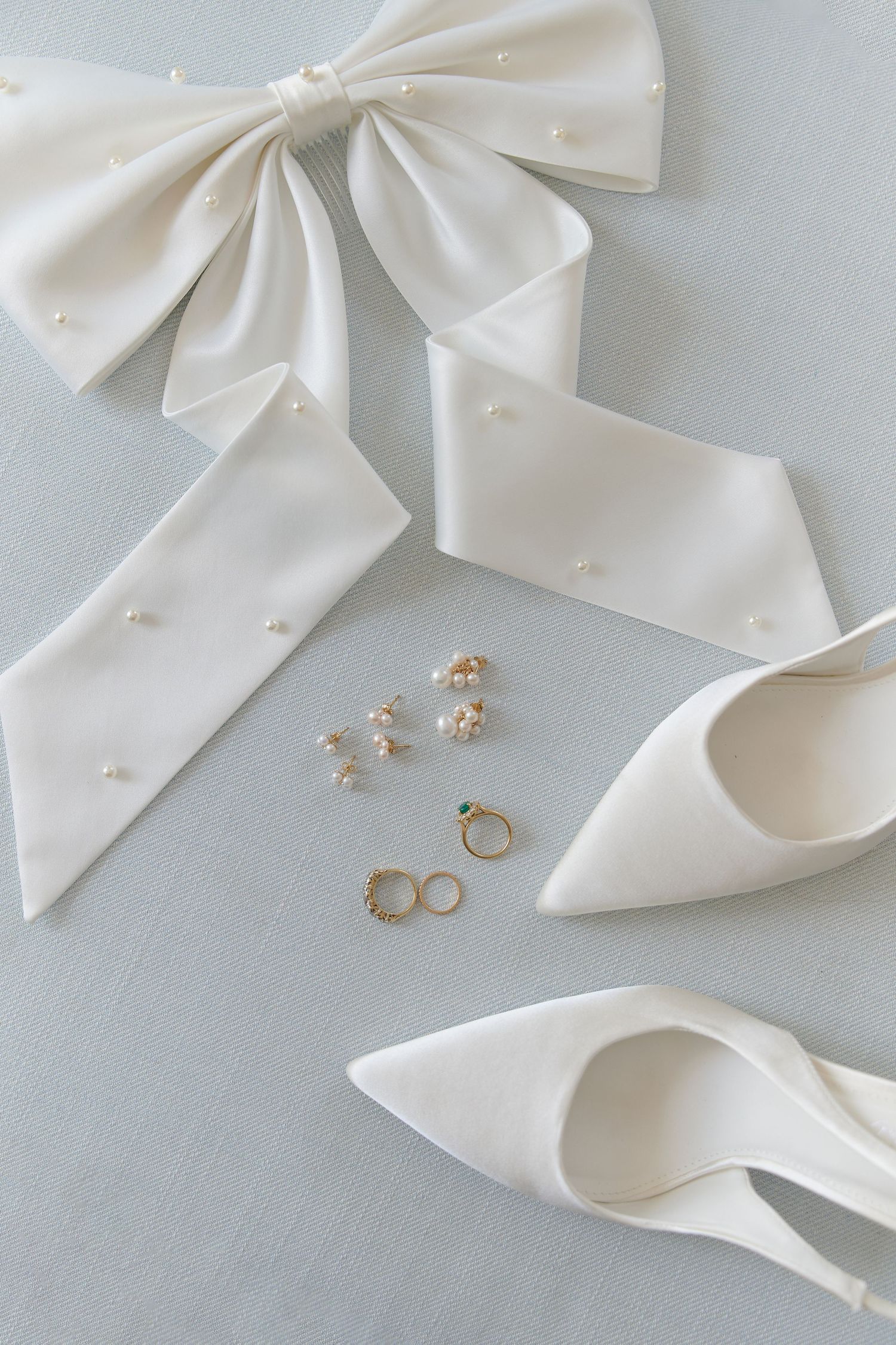White satin bridal bow, pearl earrings, gold rings, and pointed-toe heels flatlay.