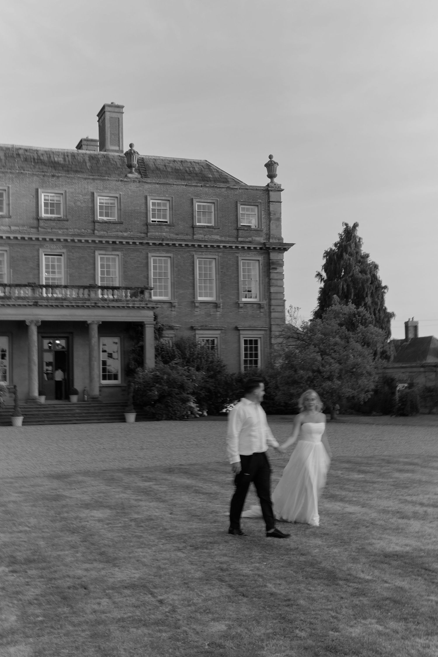 Black and white photo of a bride and groom holding hands on a lawn before a grand Georgian manor house.