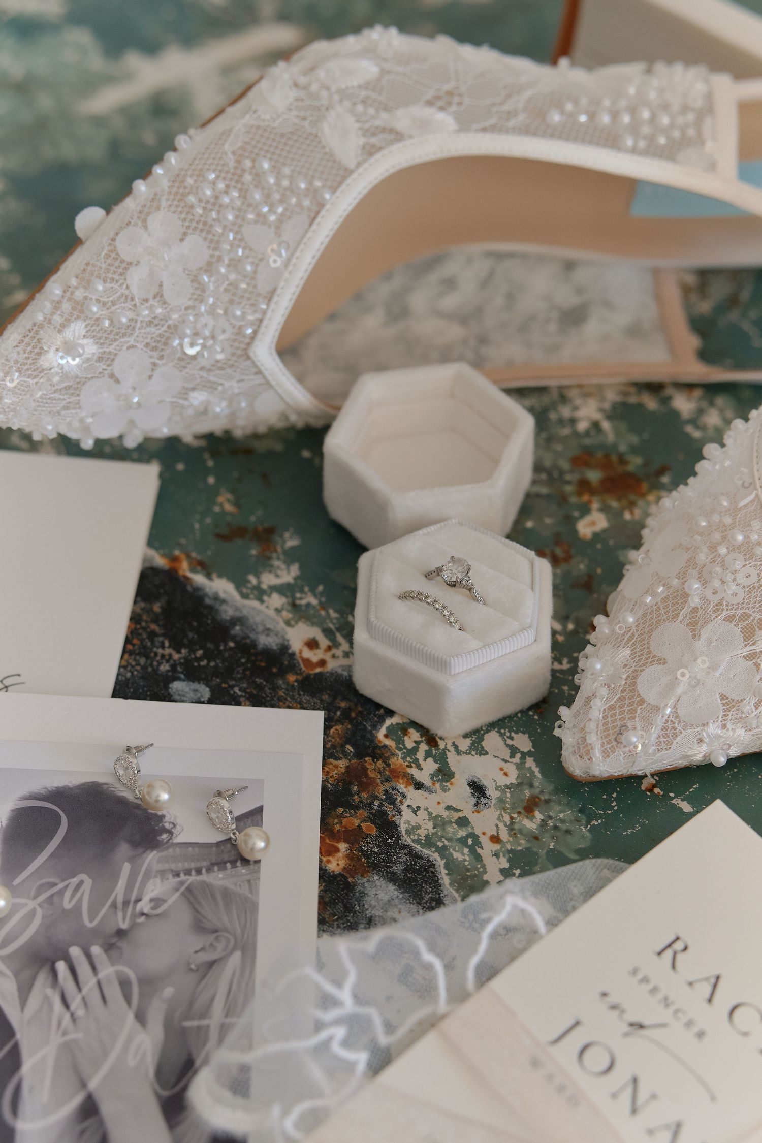 Elegant wedding flat lay with lace bridal shoes, diamond ring in velvet box, pearl earrings, and invitation suite.