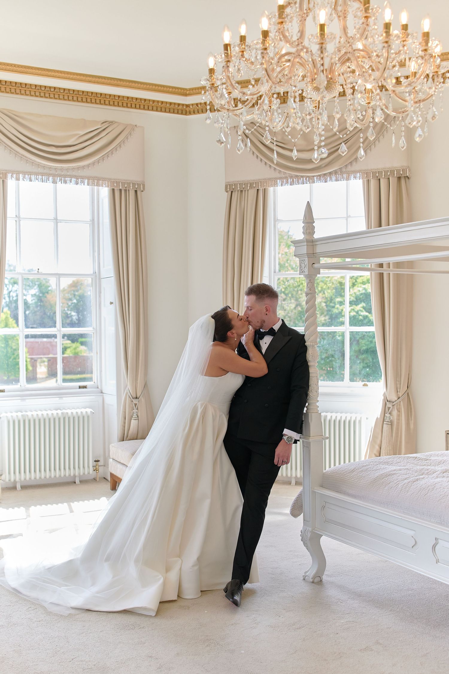 Bride and groom share a romantic kiss in an elegant room with crystal chandelier and four-poster bed.