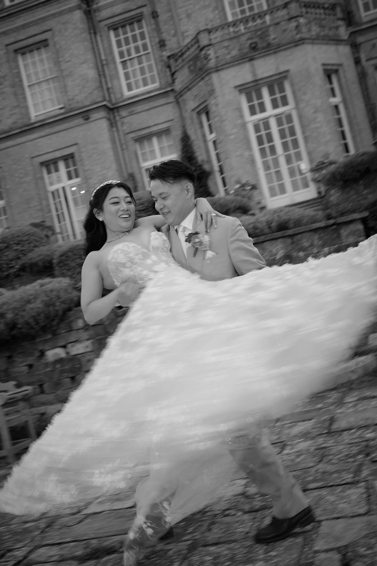 Black and white photo of a joyful bride and groom dancing outside a grand estate, her wedding dress flowing in the wind.