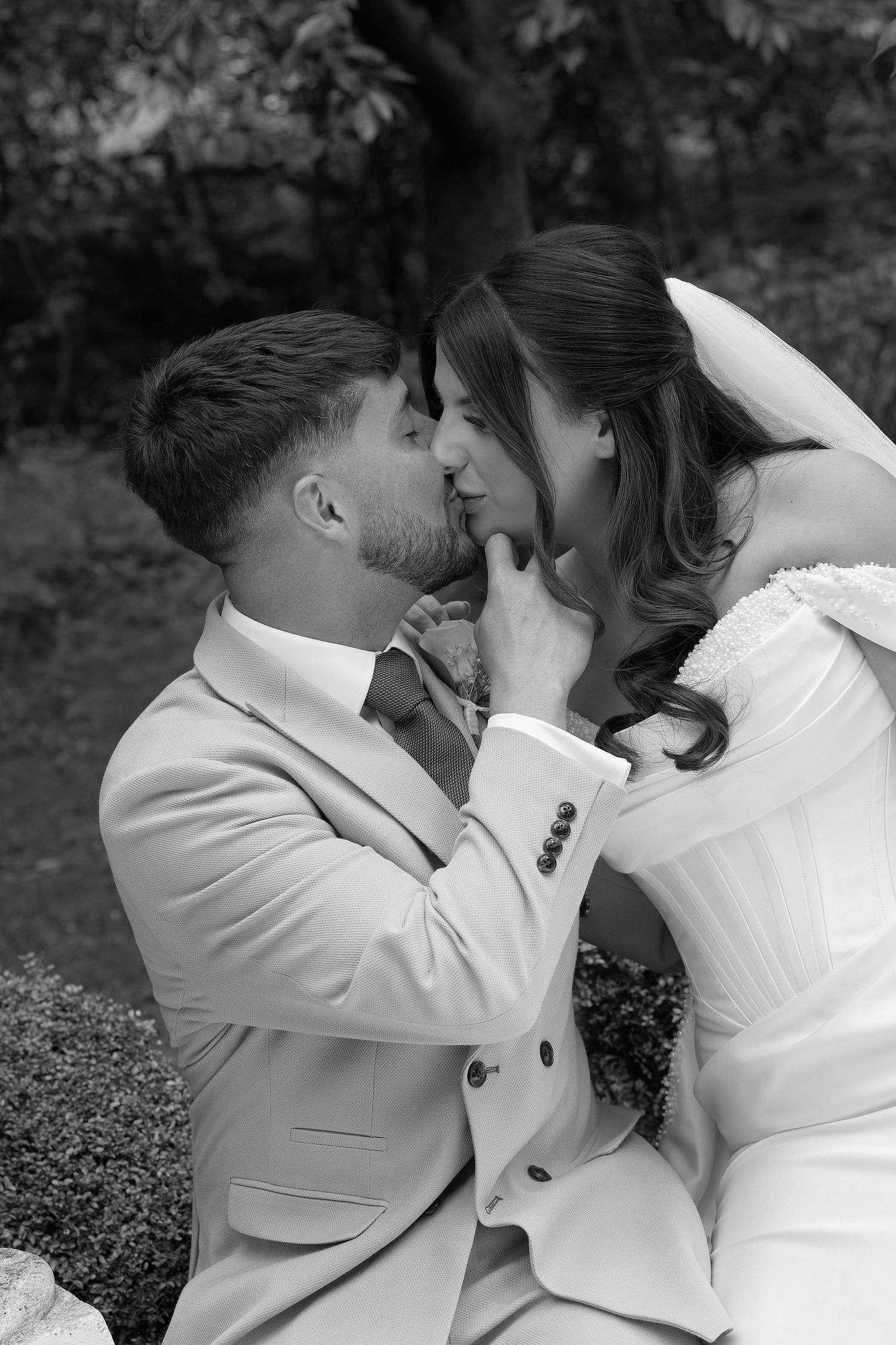Black and white photo of a groom in a suit tenderly kissing his bride in a wedding dress outdoors.