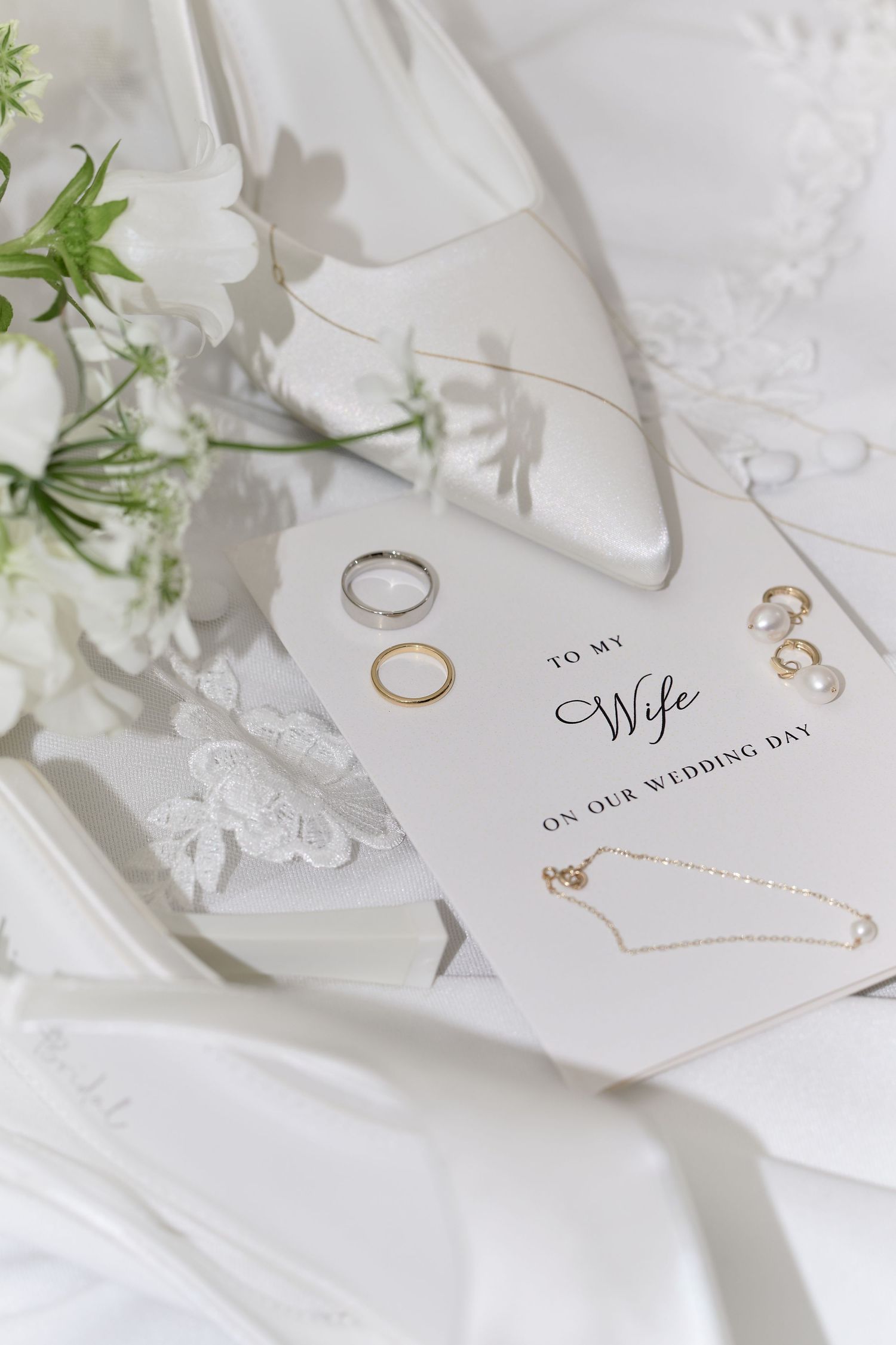 Elegant wedding flat lay with gold rings, pearl earrings, necklace, and a To My Wife card on white satin.