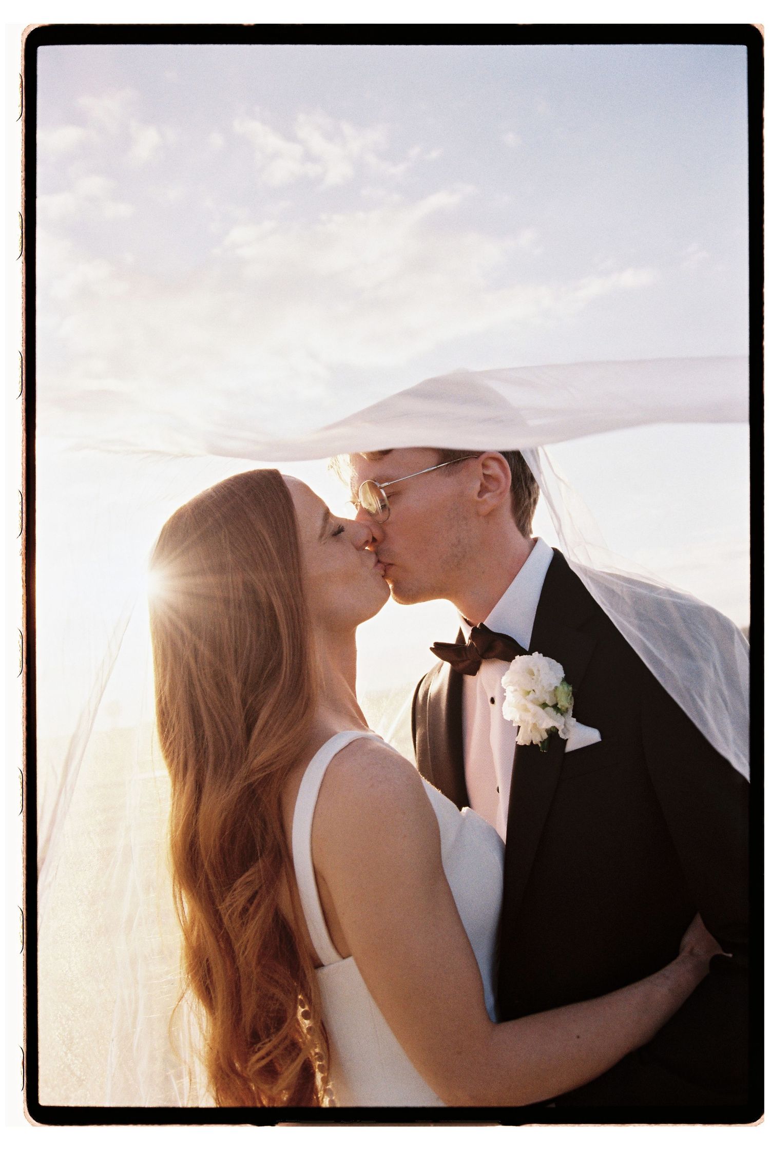 Wedding couple kissing under flowing veil at golden sunset with lens flare.