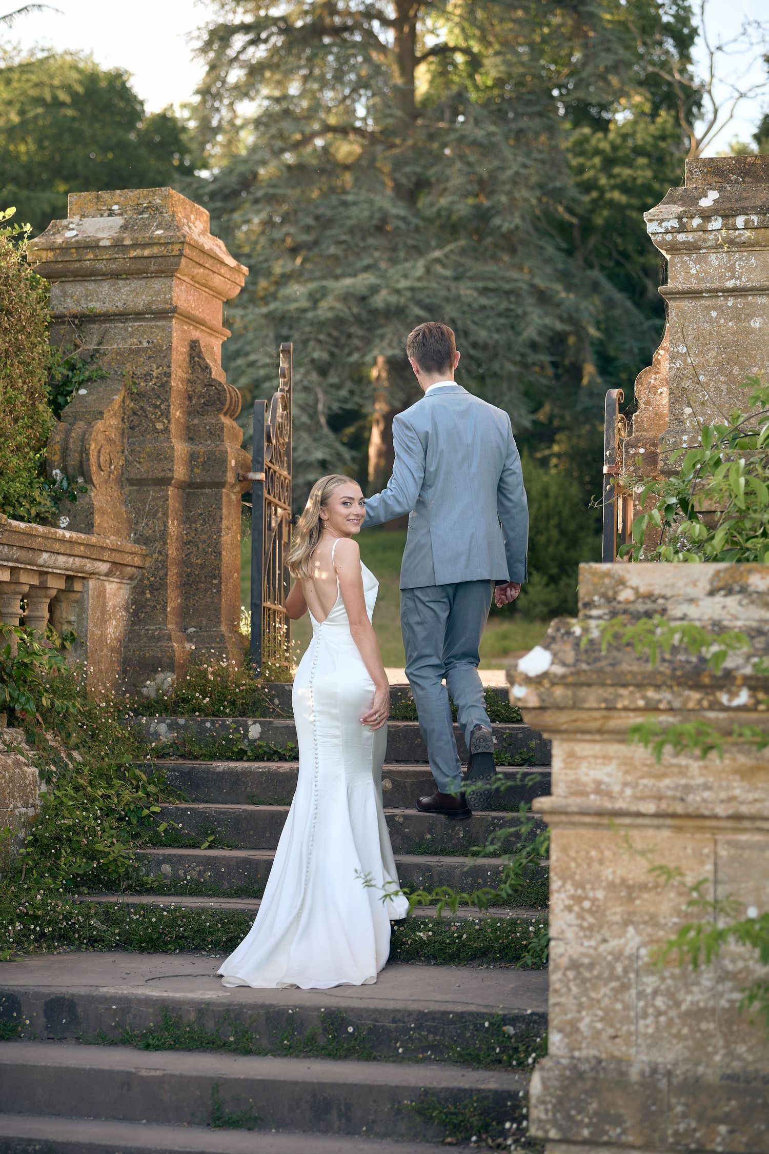 Bride in white gown looks back while holding groom's hand ascending stone steps in a garden.