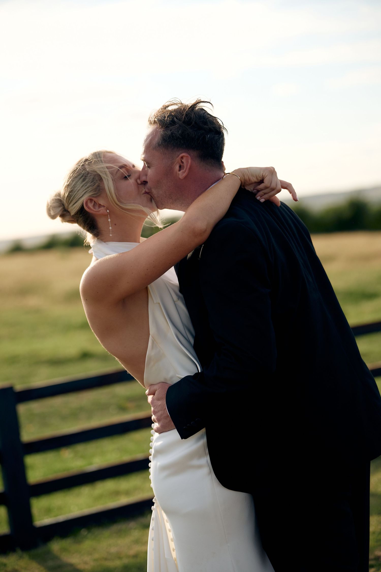 Bride and groom sharing a romantic kiss outdoors in a green field, bride in white gown, groom in dark suit.