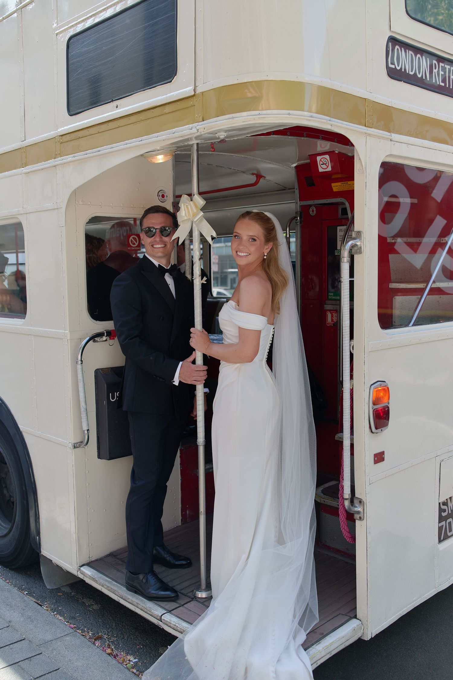 Bride and groom smiling on a vintage London double-decker wedding bus, bride in white gown with veil.