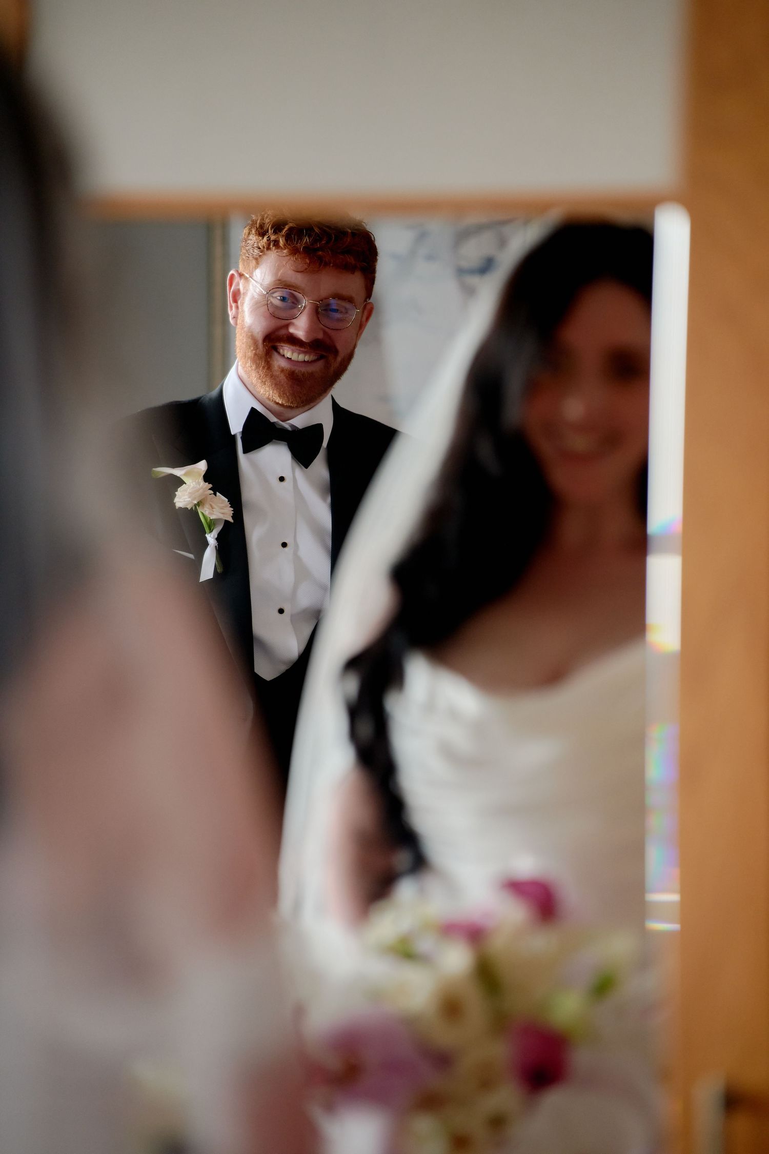 Groom in black tuxedo smiling joyfully at bride in white gown holding floral bouquet, reflected in mirror.