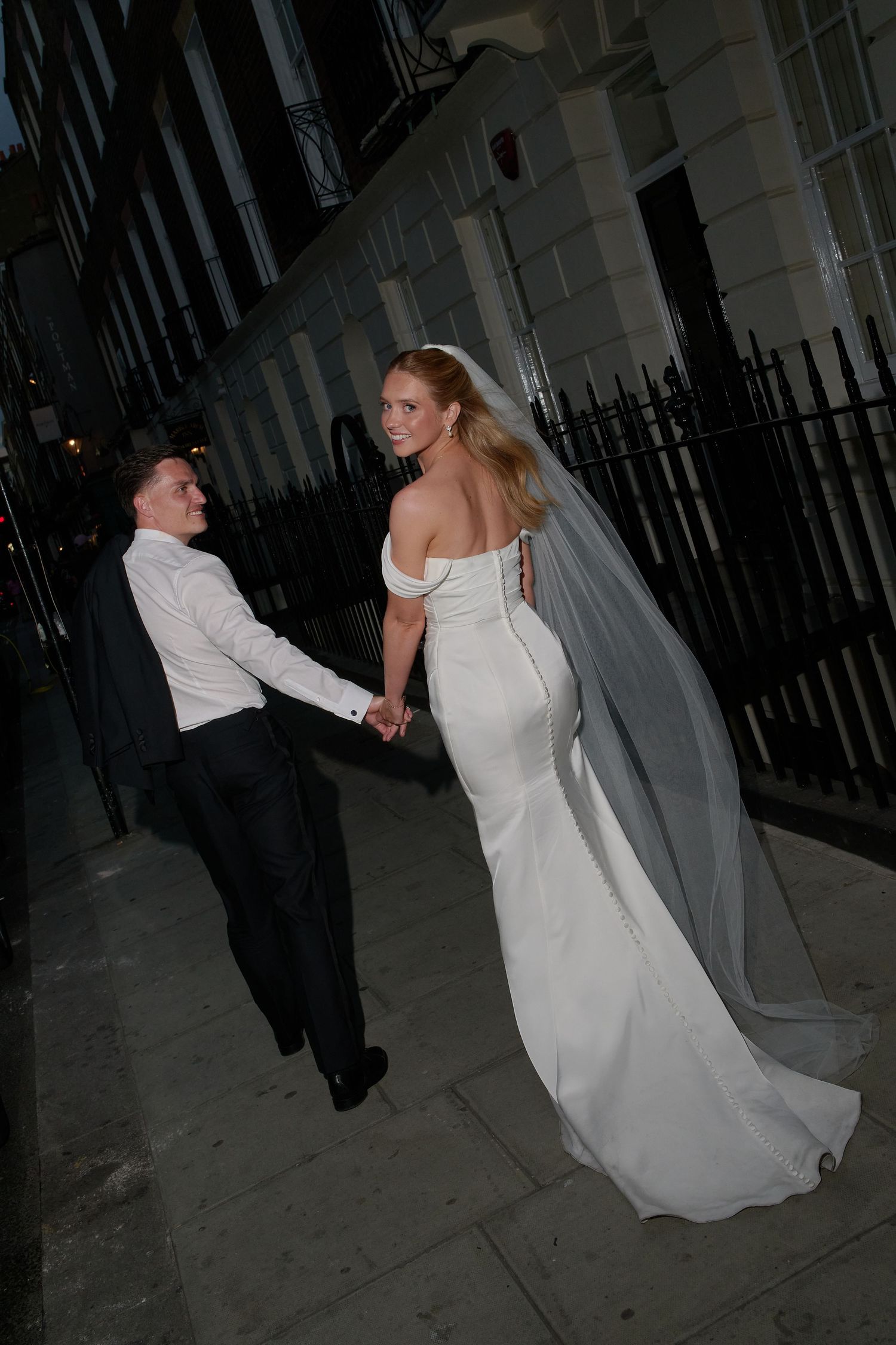 Bride in white off-shoulder gown with veil holds groom's hand walking along evening city street.