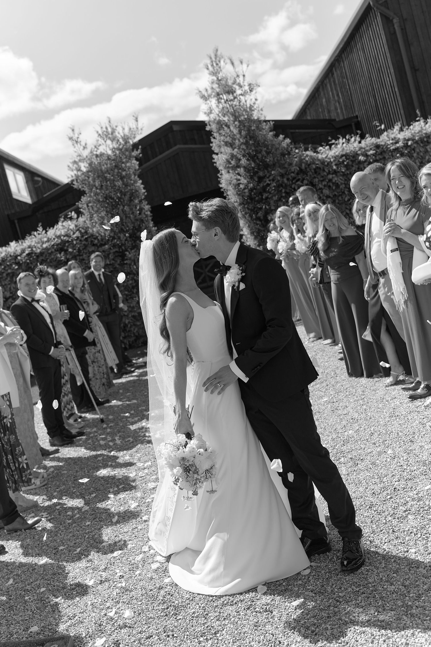 Black and white photo of a bride and groom sharing a kiss while guests line the path throwing confetti.
