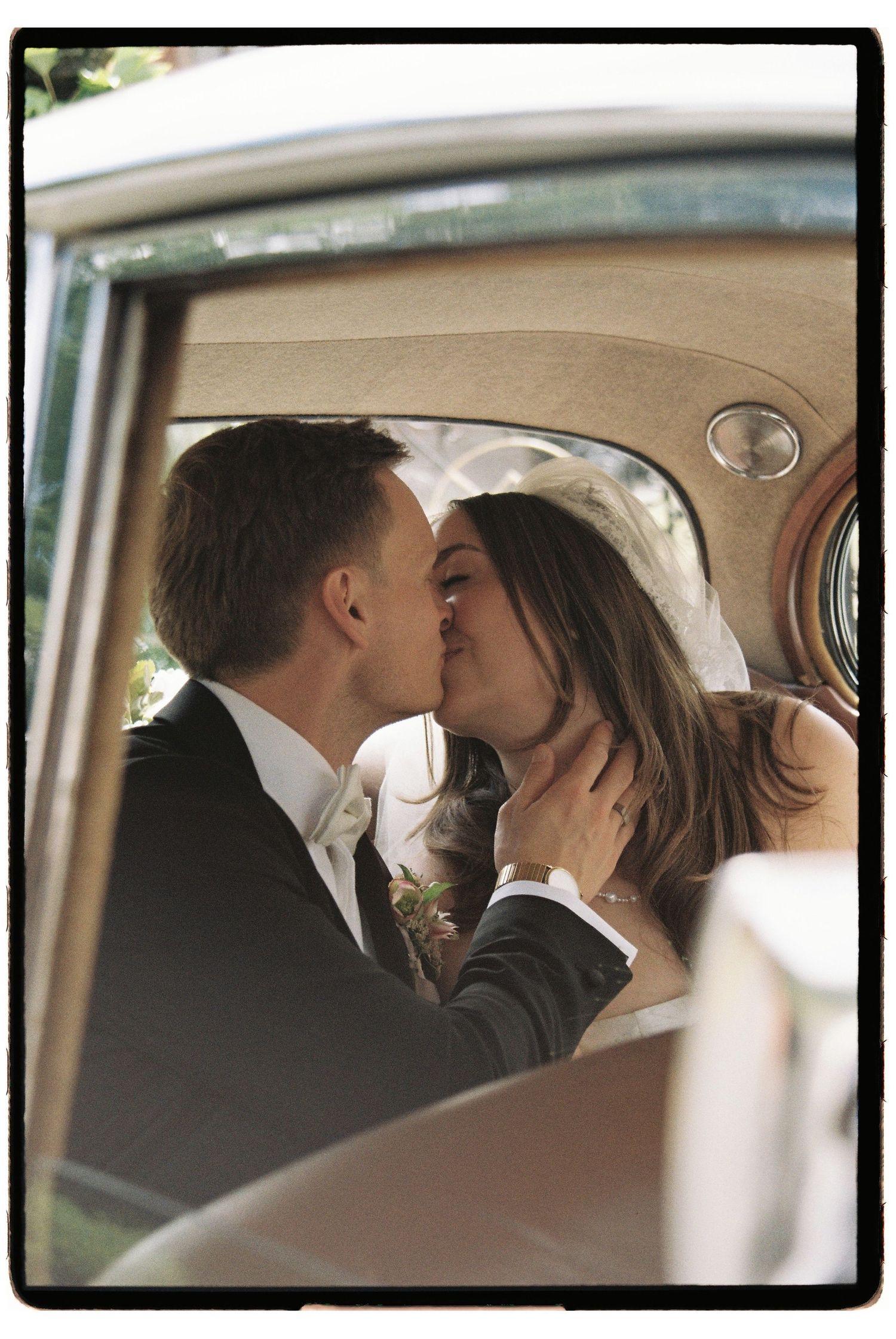 Newlywed couple sharing a romantic kiss inside a vintage car on their wedding day.