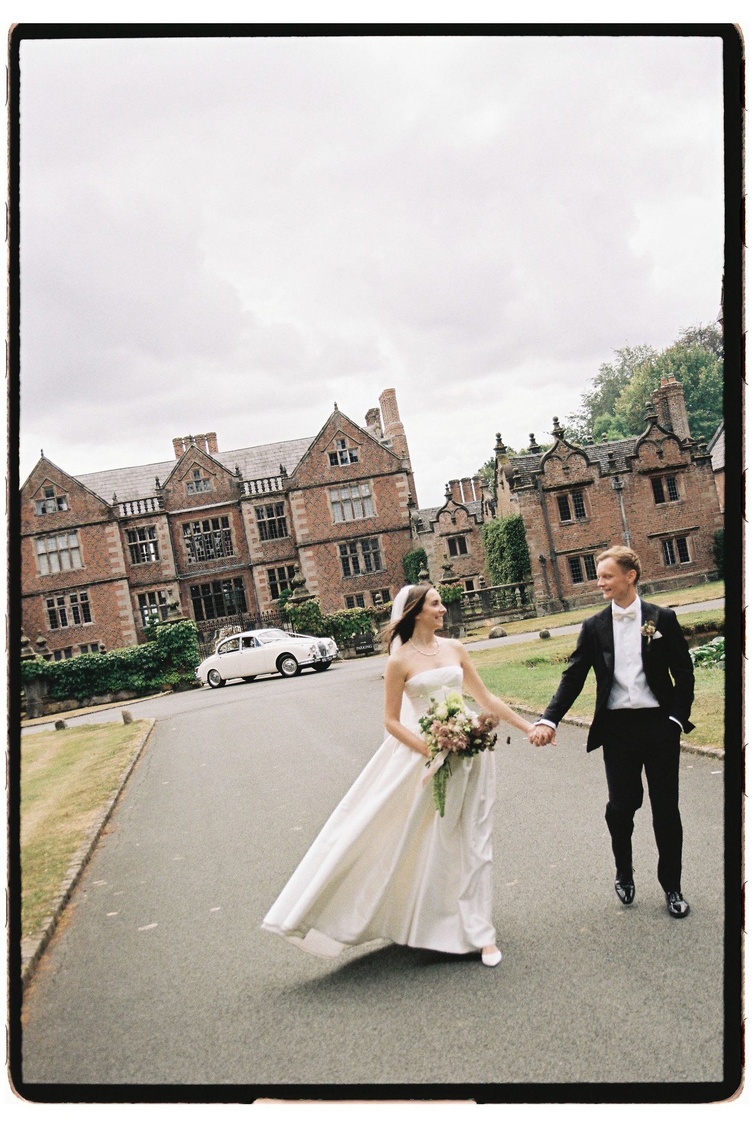 Bride and groom walk hand in hand outside a grand historic manor house on their wedding day.