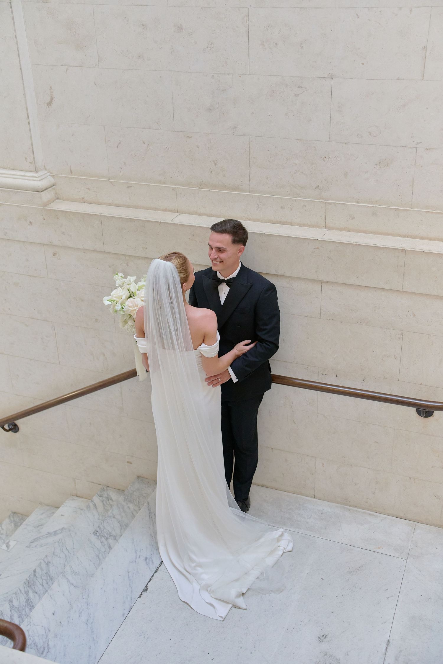 Bride and groom share a romantic moment on marble stairs, with bride in white gown and veil holding bouquet.
