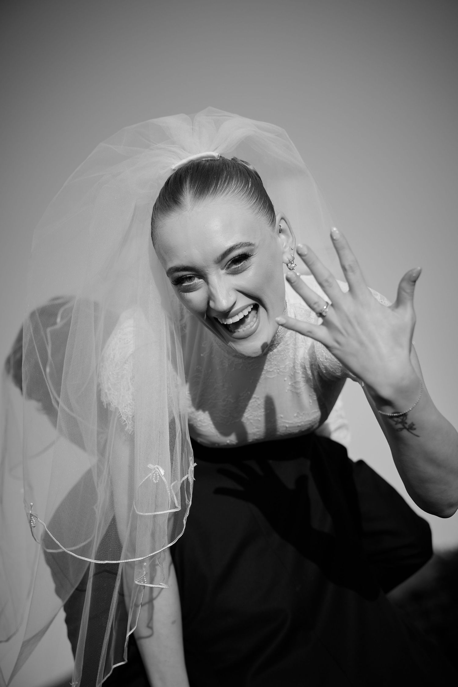 Joyful bride in veil showing engagement ring, laughing in black and white portrait photo.