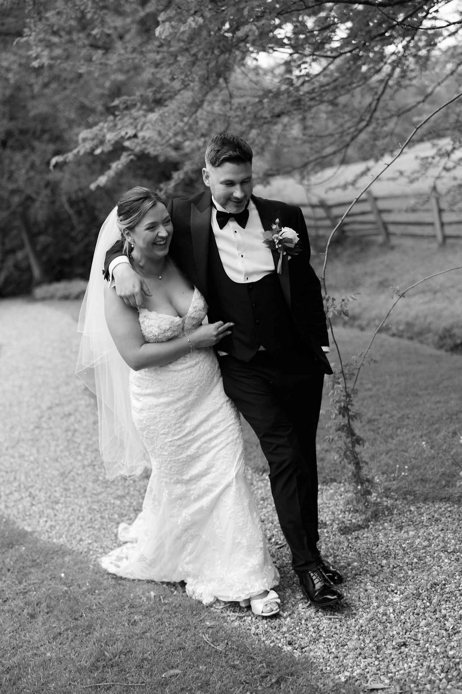 Black and white photo of a smiling bride in lace gown and groom in tuxedo walking together outdoors on their wedding day.