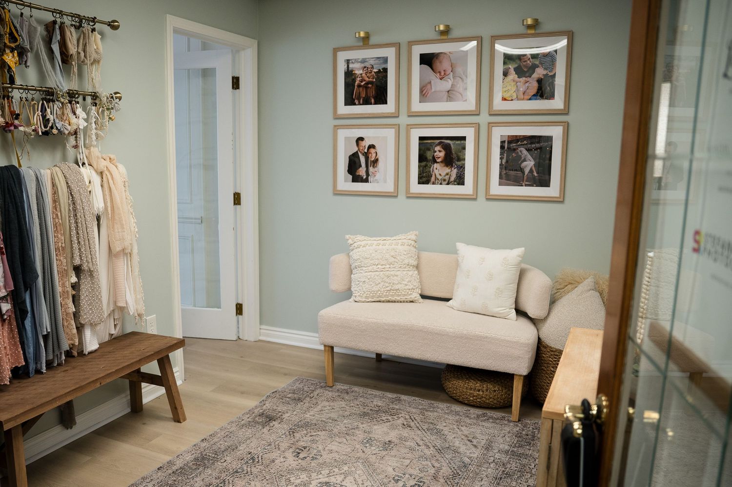 Elegant boutique sitting area with cream chaise lounge, gallery wall of framed photos, and clothing rack on sage green walls.