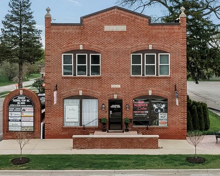 Red brick two-story commercial building with signage, arched windows, and landscaped entrance on a sunny day.