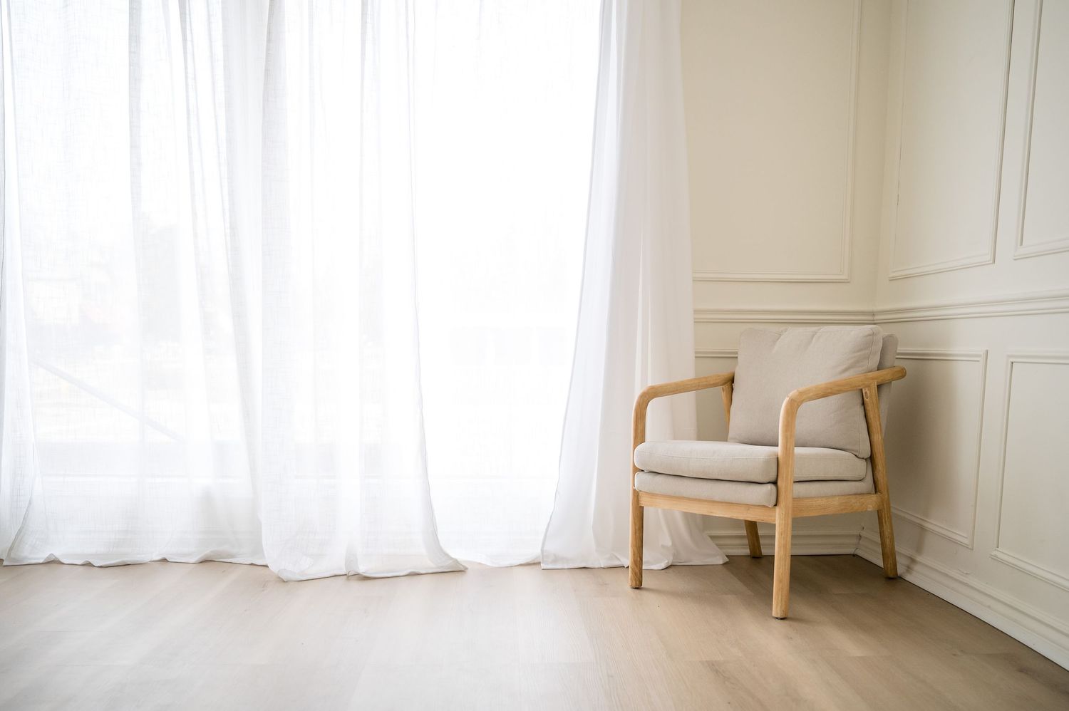 A minimalist wooden armchair with beige cushions sits in a bright room with white curtains and light hardwood floors.