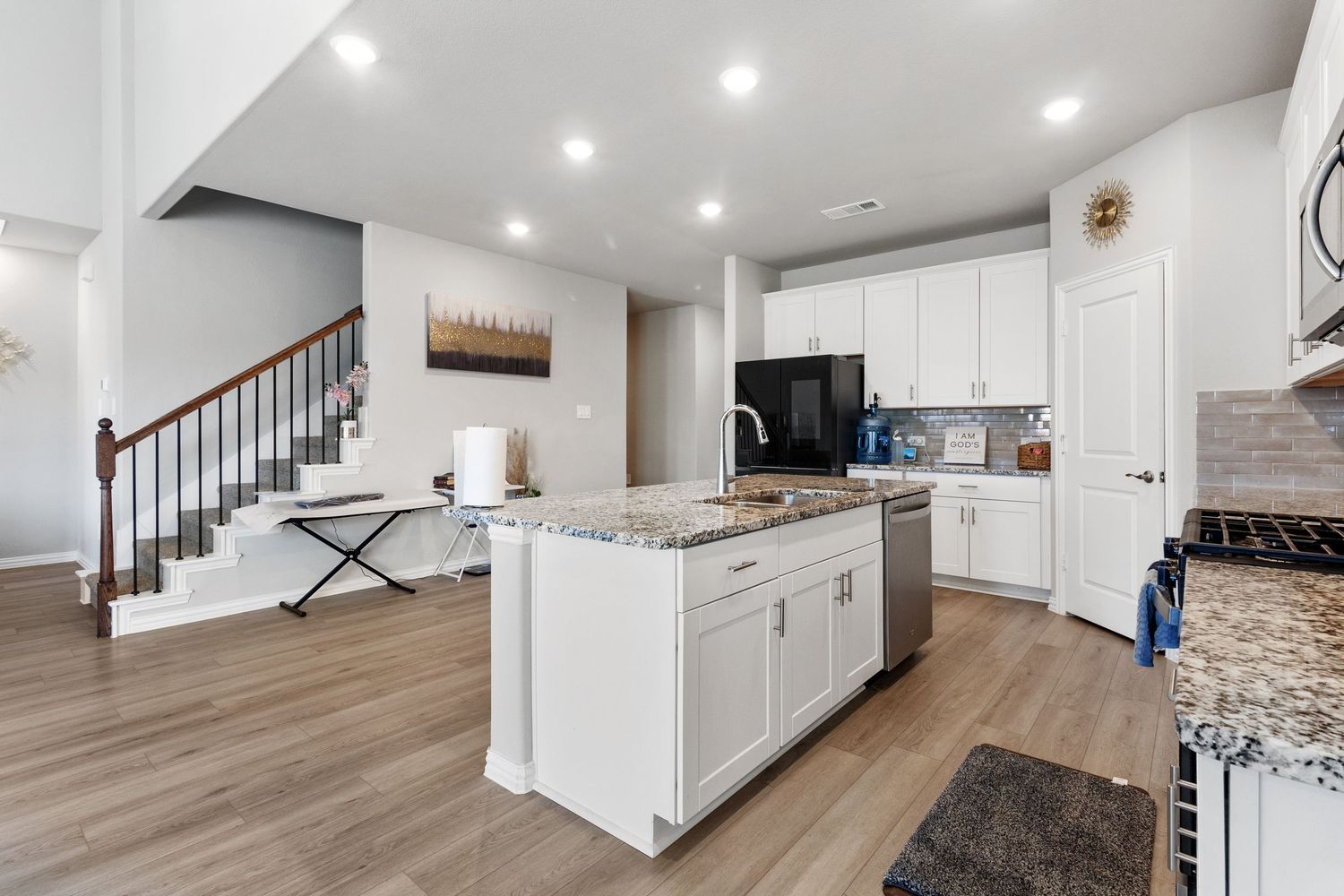 Modern kitchen with white cabinets, granite island, black appliances, hardwood floors, and open staircase in background.
