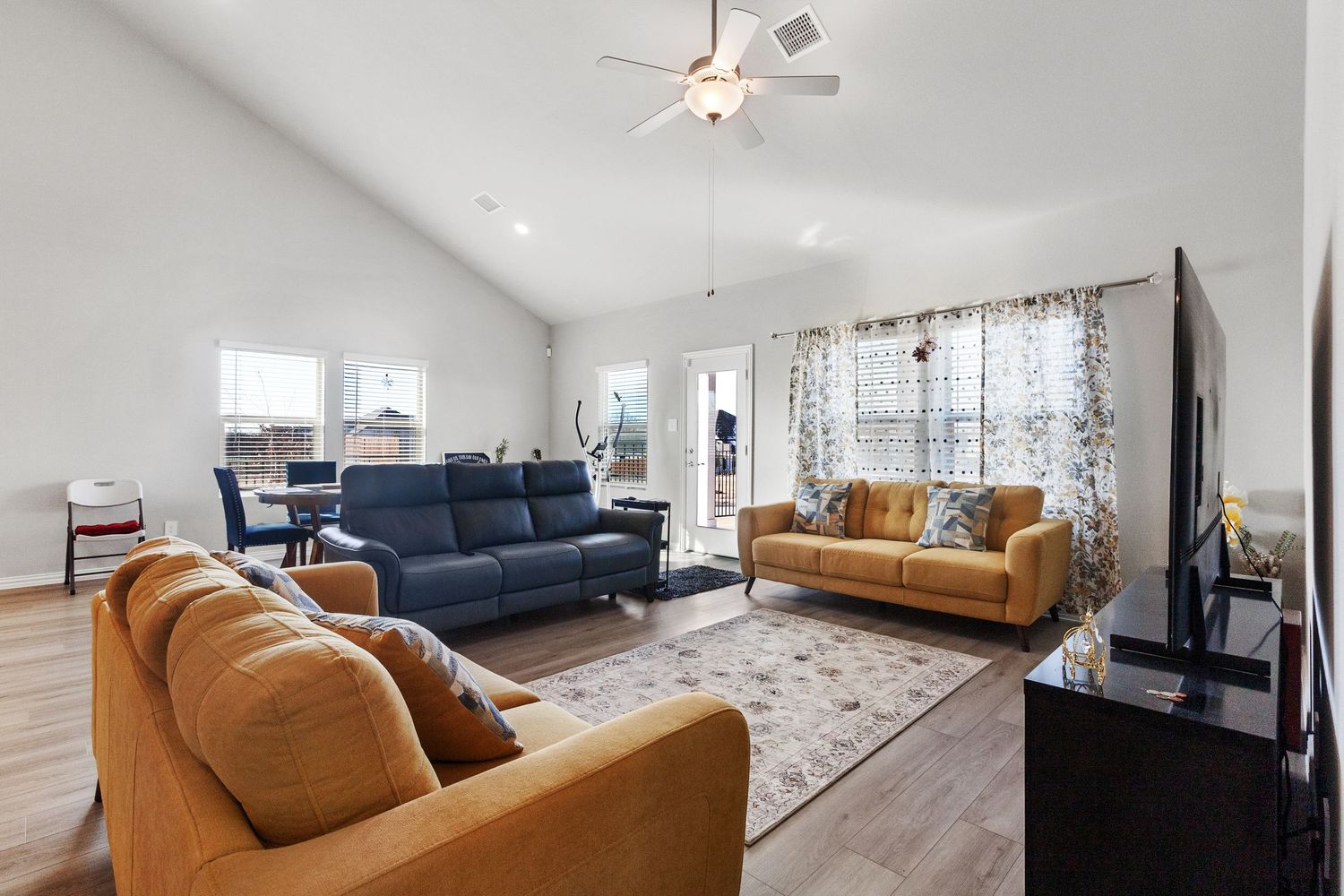 Bright living room with vaulted ceiling, ceiling fan, tan and blue sofas on a patterned rug, and hardwood floors.
