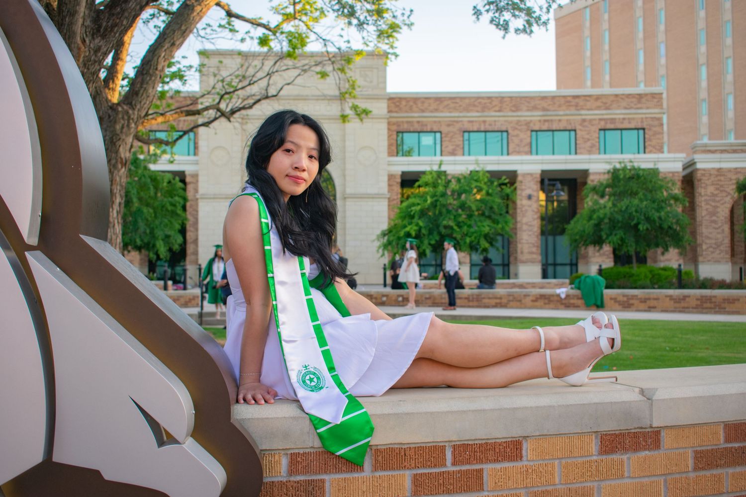 Young woman in white dress and green graduation sash poses on brick wall at university campus.
