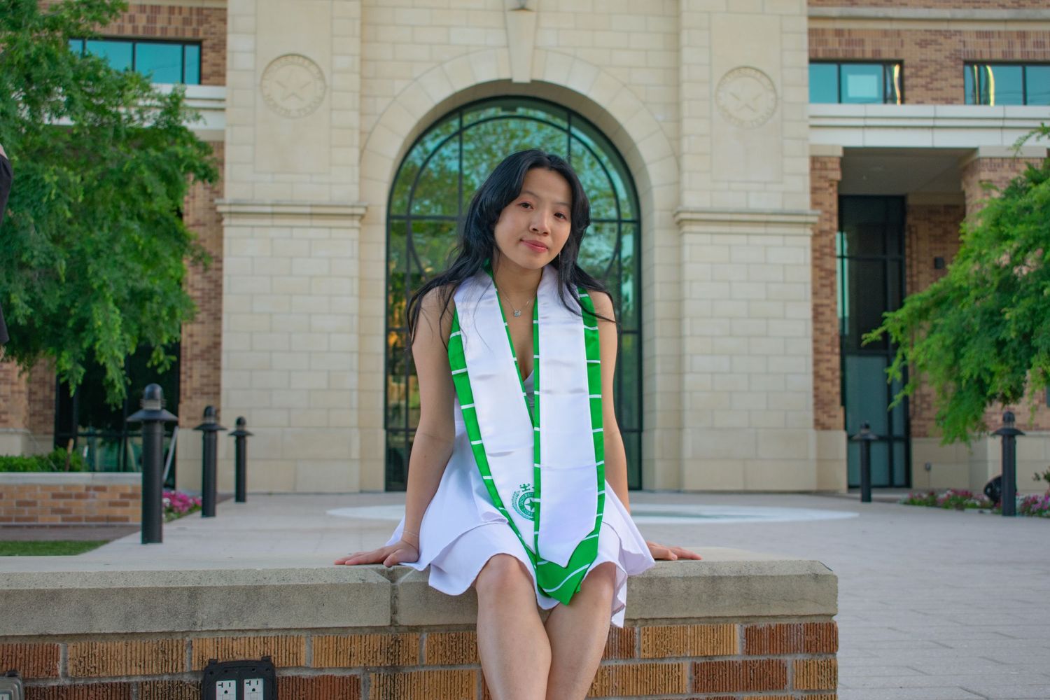 Young woman in white dress and green graduation stole sitting on brick wall outside university building.