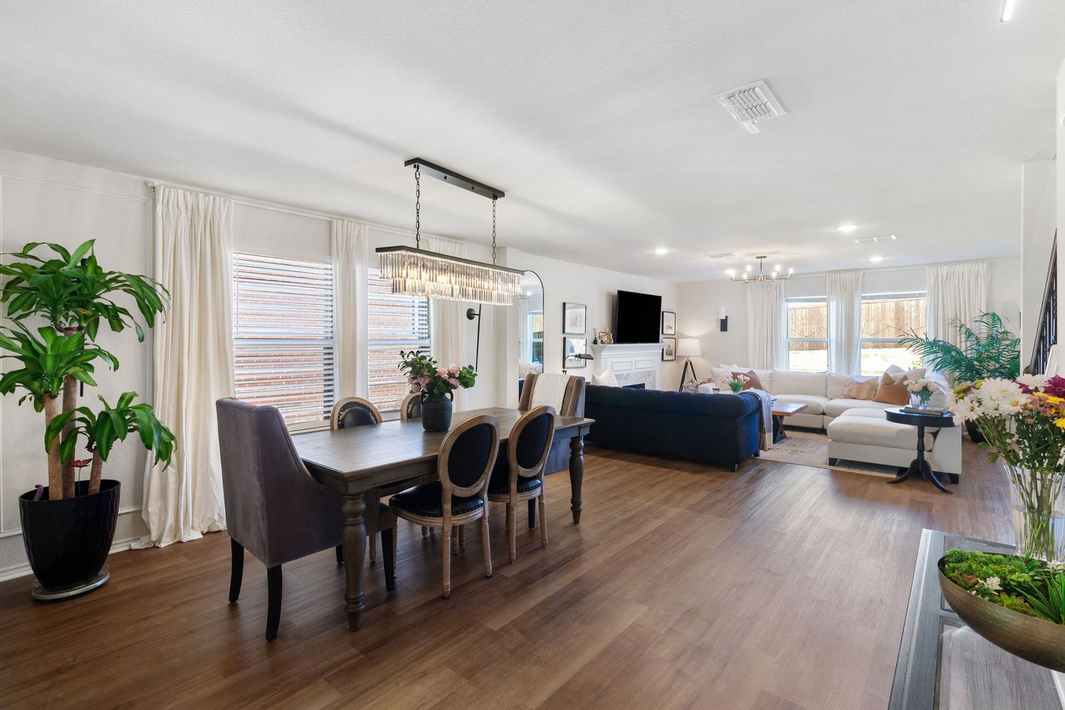 Open-concept dining and living room with dark hardwood floors, chandelier, gray chairs, and abundant natural light.