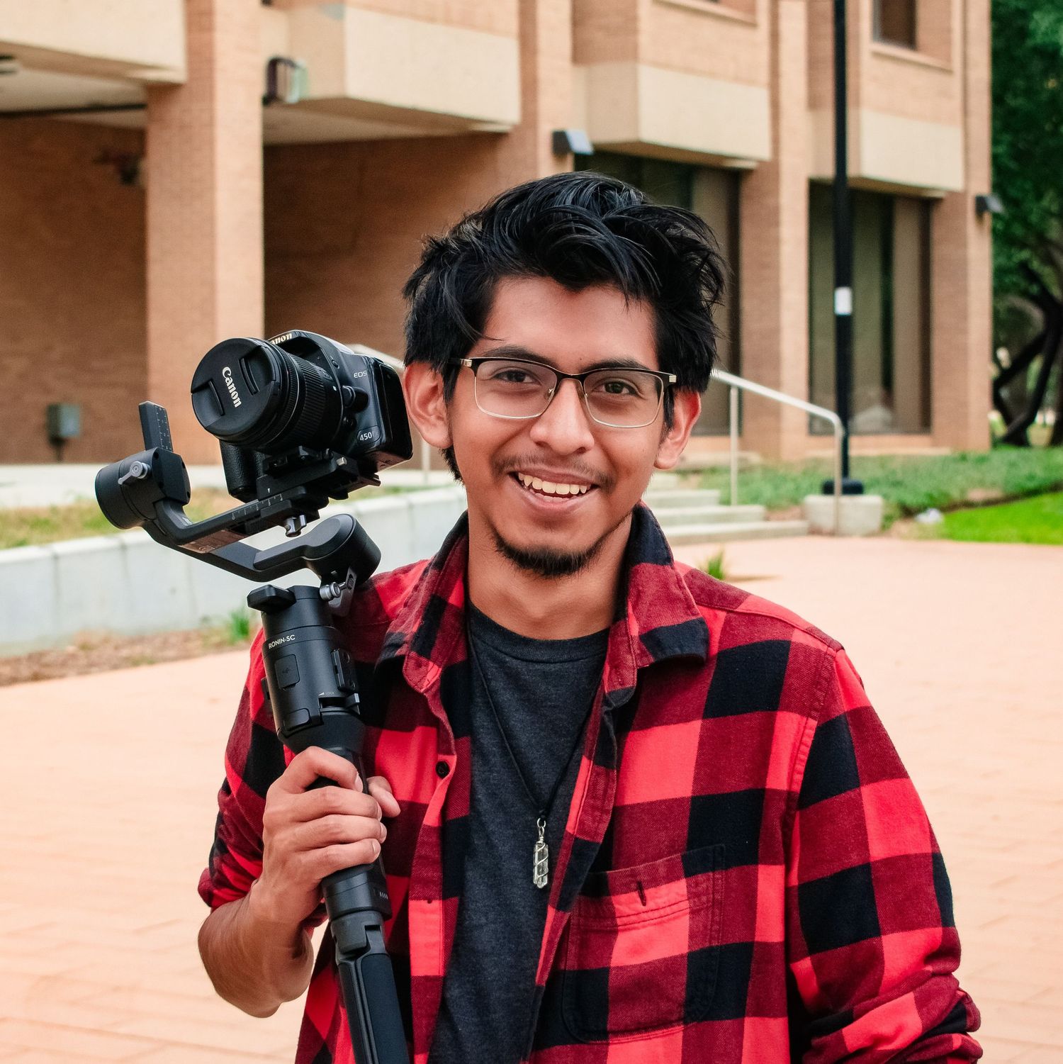 Young man smiling, holding a camera gimbal stabilizer, wearing a red plaid shirt outdoors on a campus.