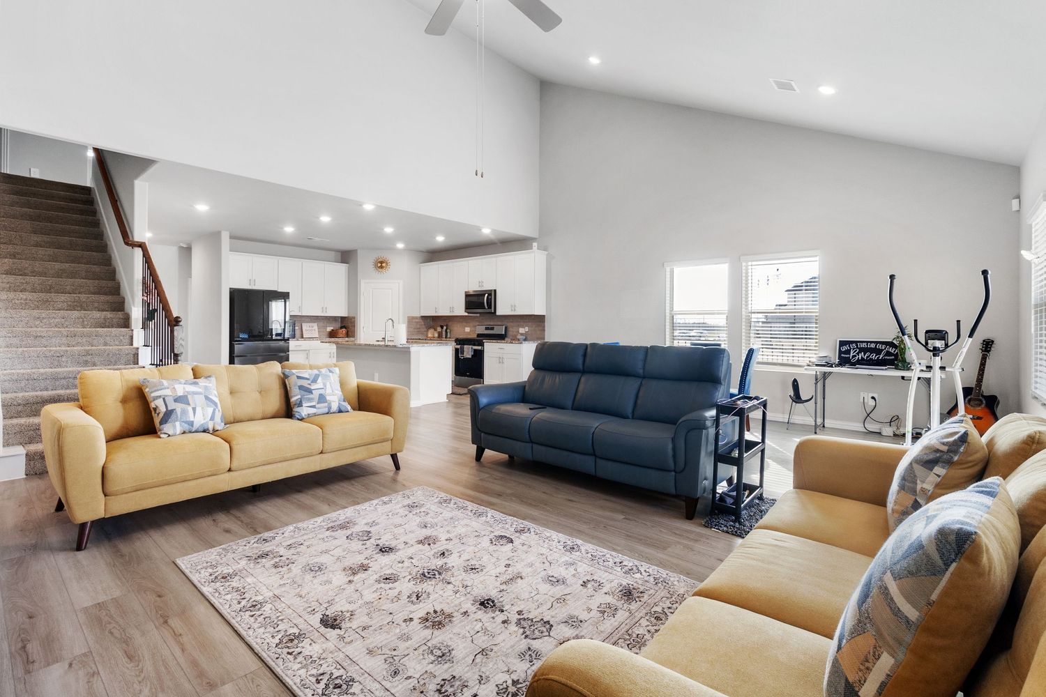 Open-concept living room with vaulted ceilings, yellow sofas, blue couch, hardwood floors, and modern kitchen in background.