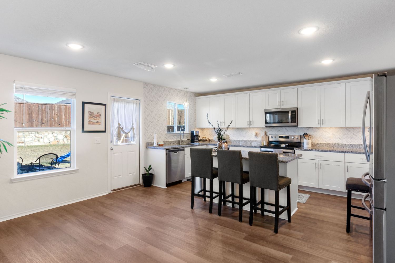Modern kitchen with white cabinets, stainless steel appliances, dark bar stools, and hardwood floors in an open-concept layout.