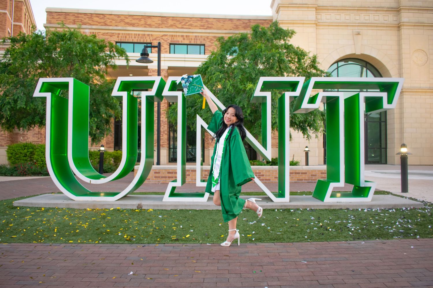 Graduate in green cap and gown poses joyfully in front of large UNT letter sign on university campus.