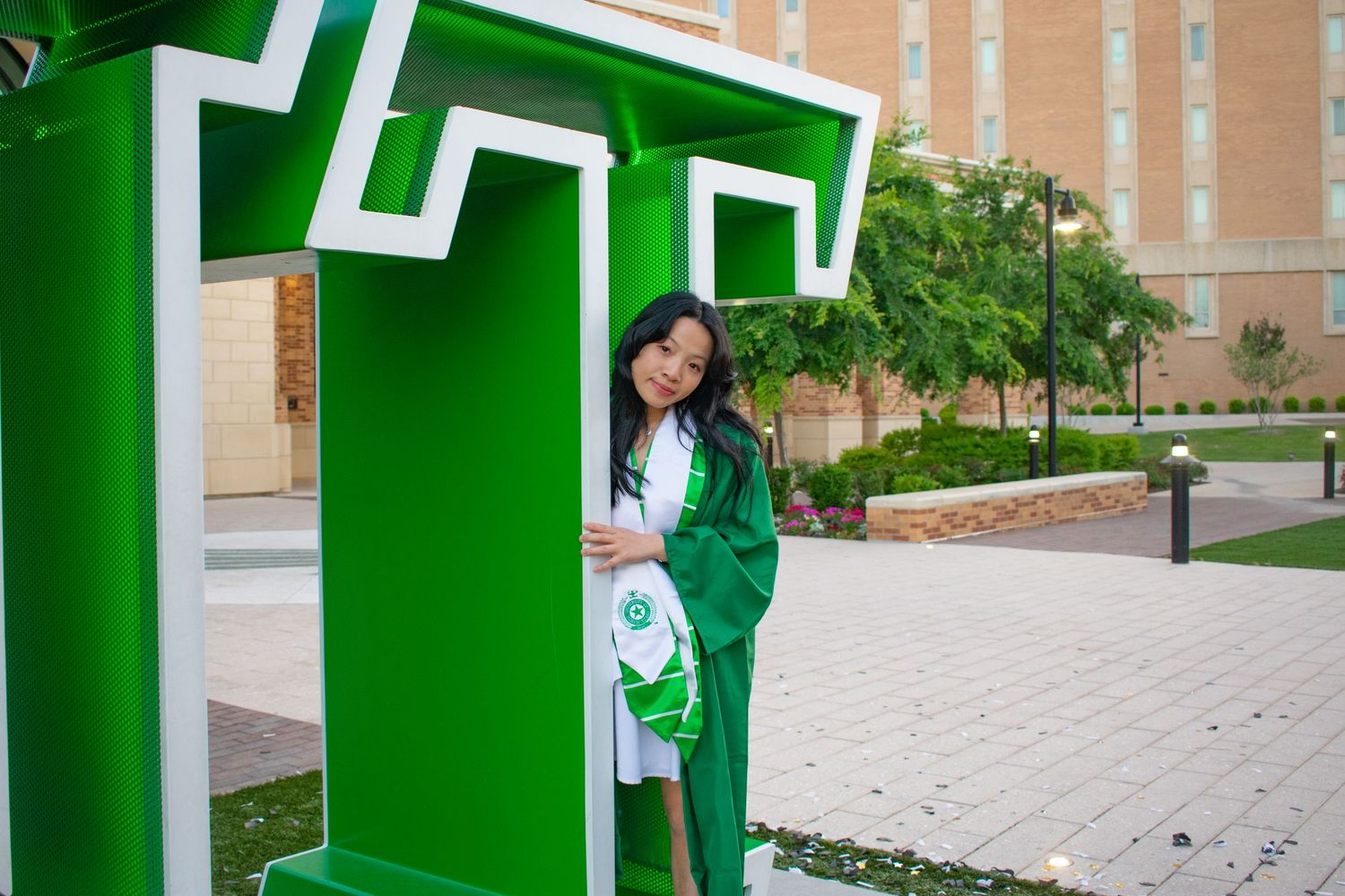 A young woman in green graduation cap and gown poses beside a large green letter T on a university campus.