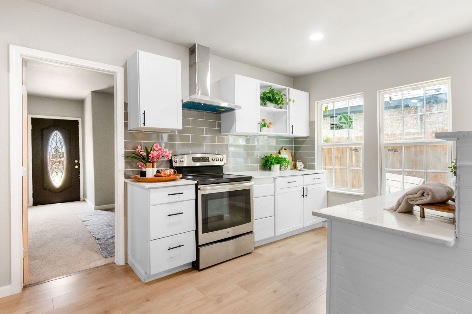 Modern white kitchen with stainless steel range, subway tile backsplash, wood floors, and bright windows overlooking a backyard.